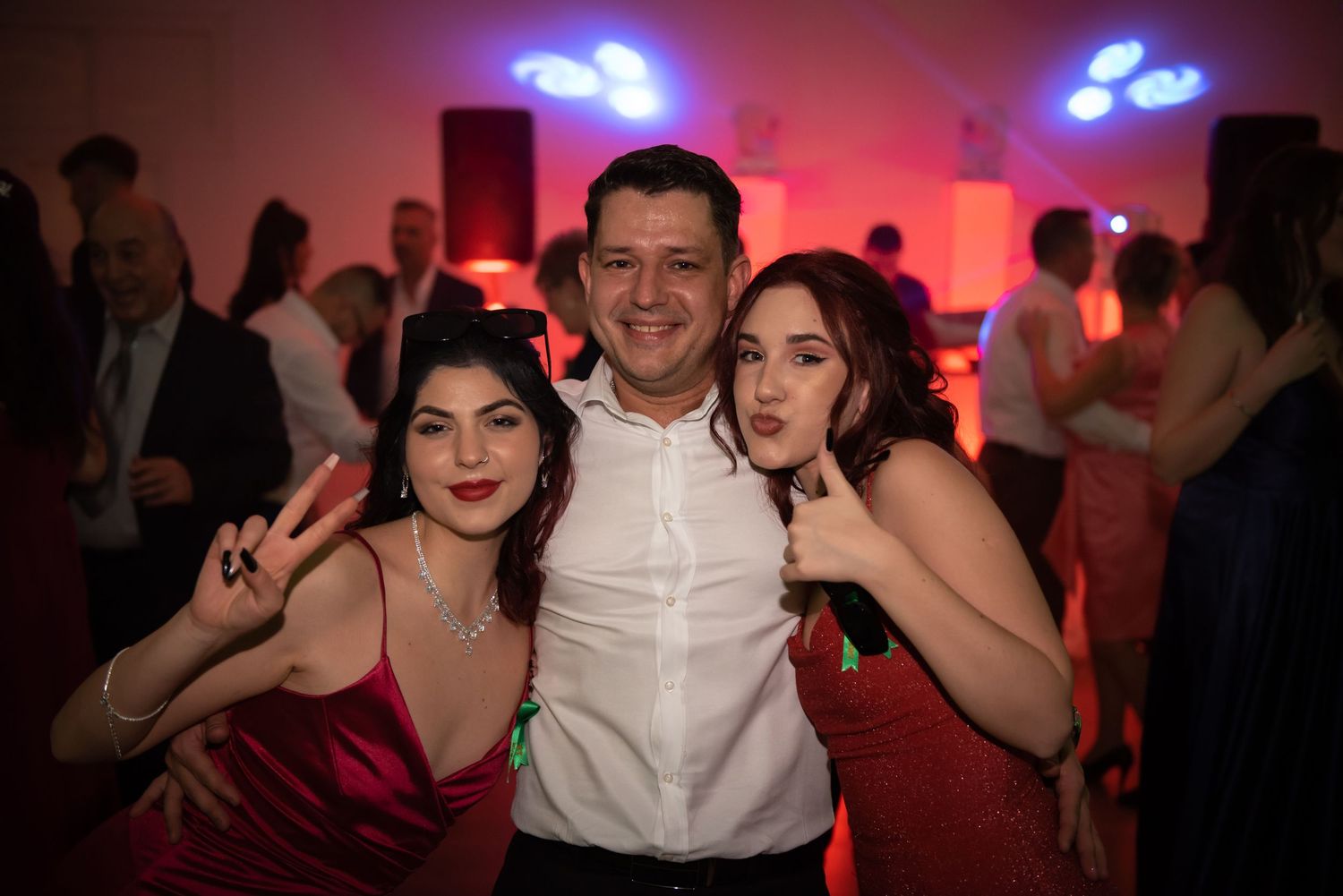 Three people smiling and posing together on the dance floor under red party lights at a high school prom.