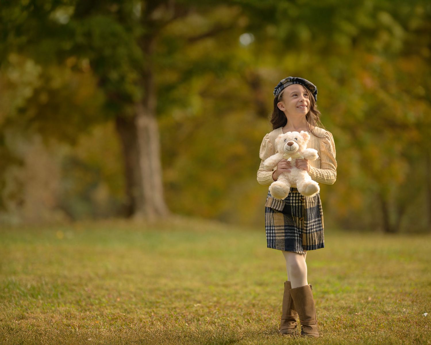 A young girl holding a white teddy bear stands in an autumn park wearing a plaid skirt and brown boots.