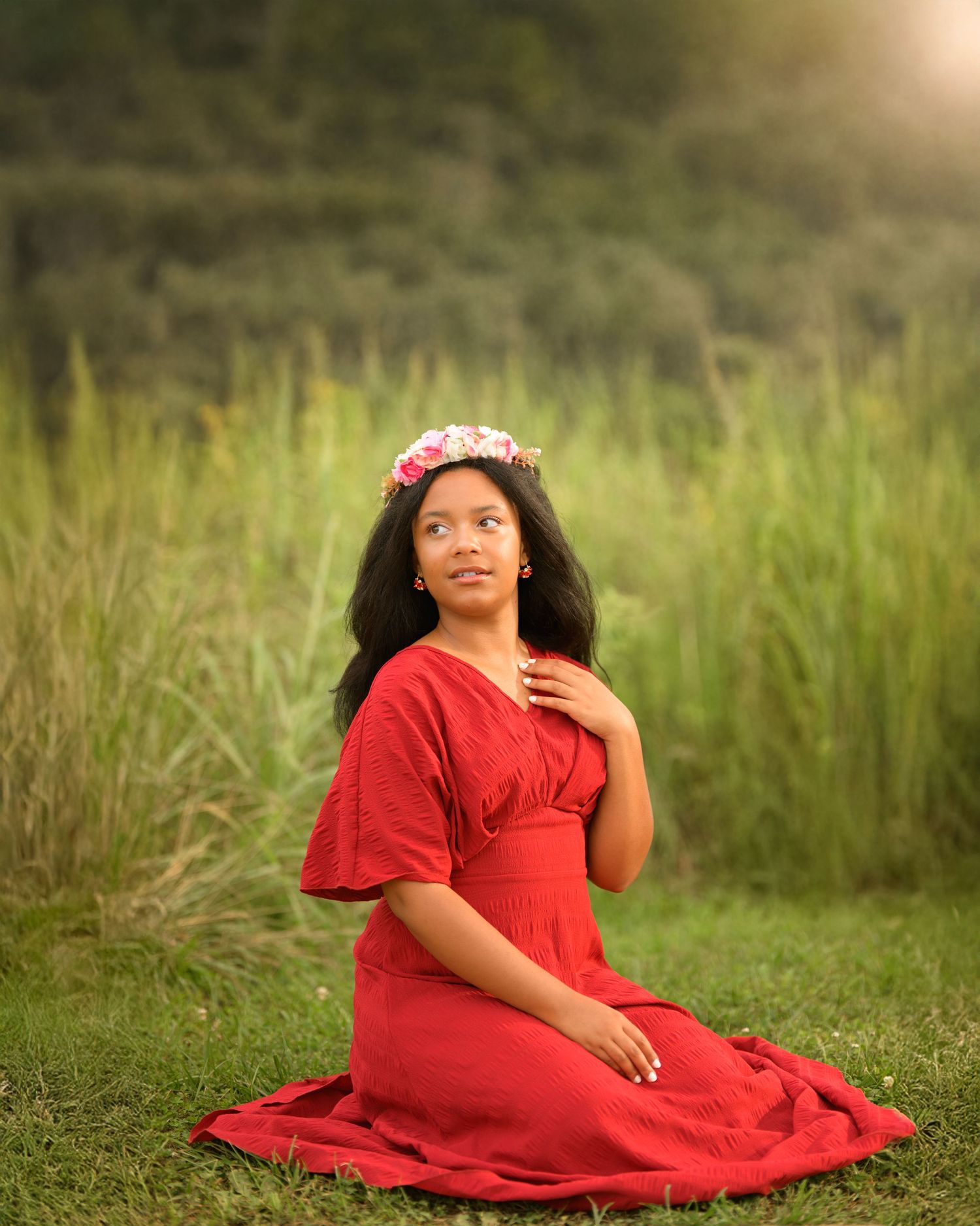 Wearing a red dress and floral crown, a person poses gracefully in a sunlit field of tall grass.