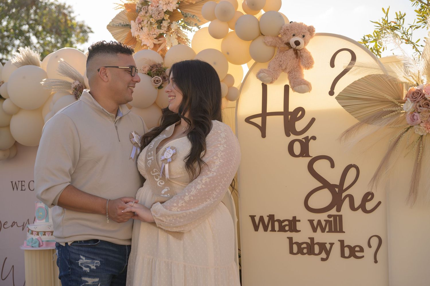 Gender reveal party setup with couple posing in front of balloon arch and decorative signage.