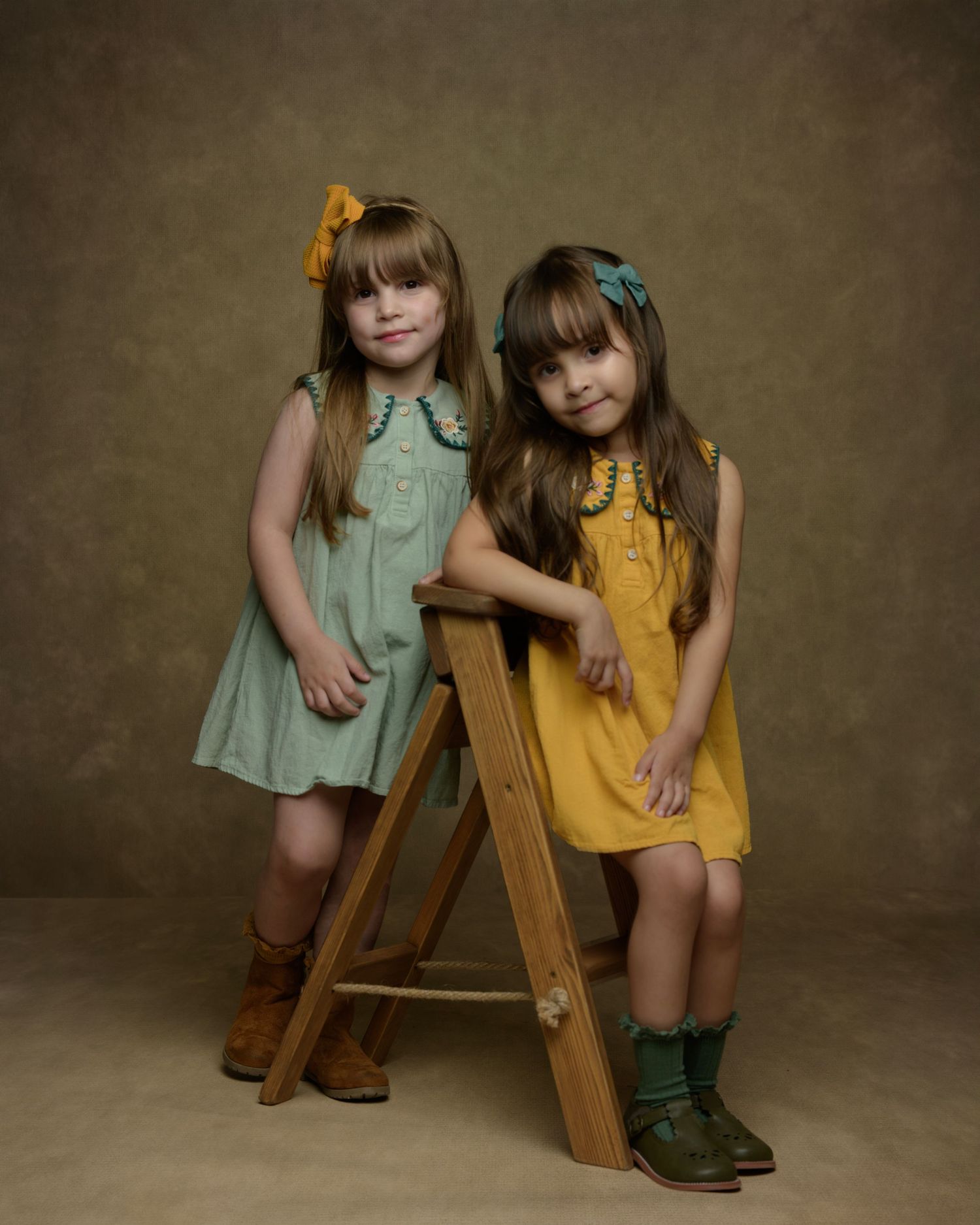 Two children in vintage-style dresses posing with wooden ladder against brown backdrop.