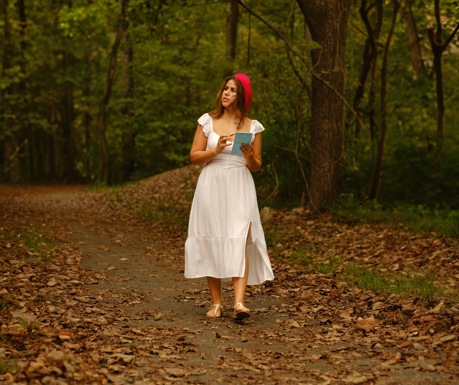 Woman in flowing white dress and red beret walking through autumn forest path.