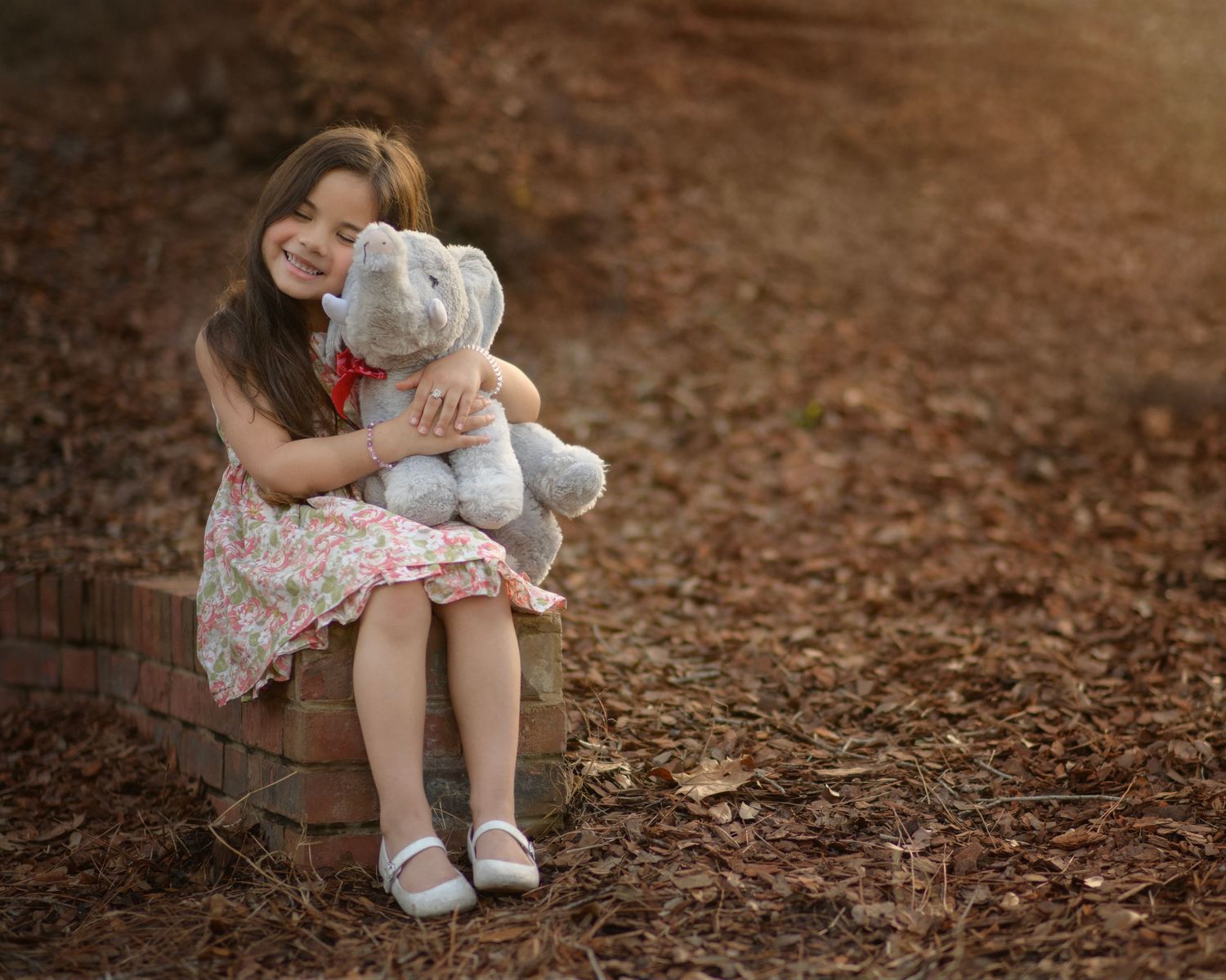 A child in a floral dress sits on brown ground hugging a plush teddy bear toy.