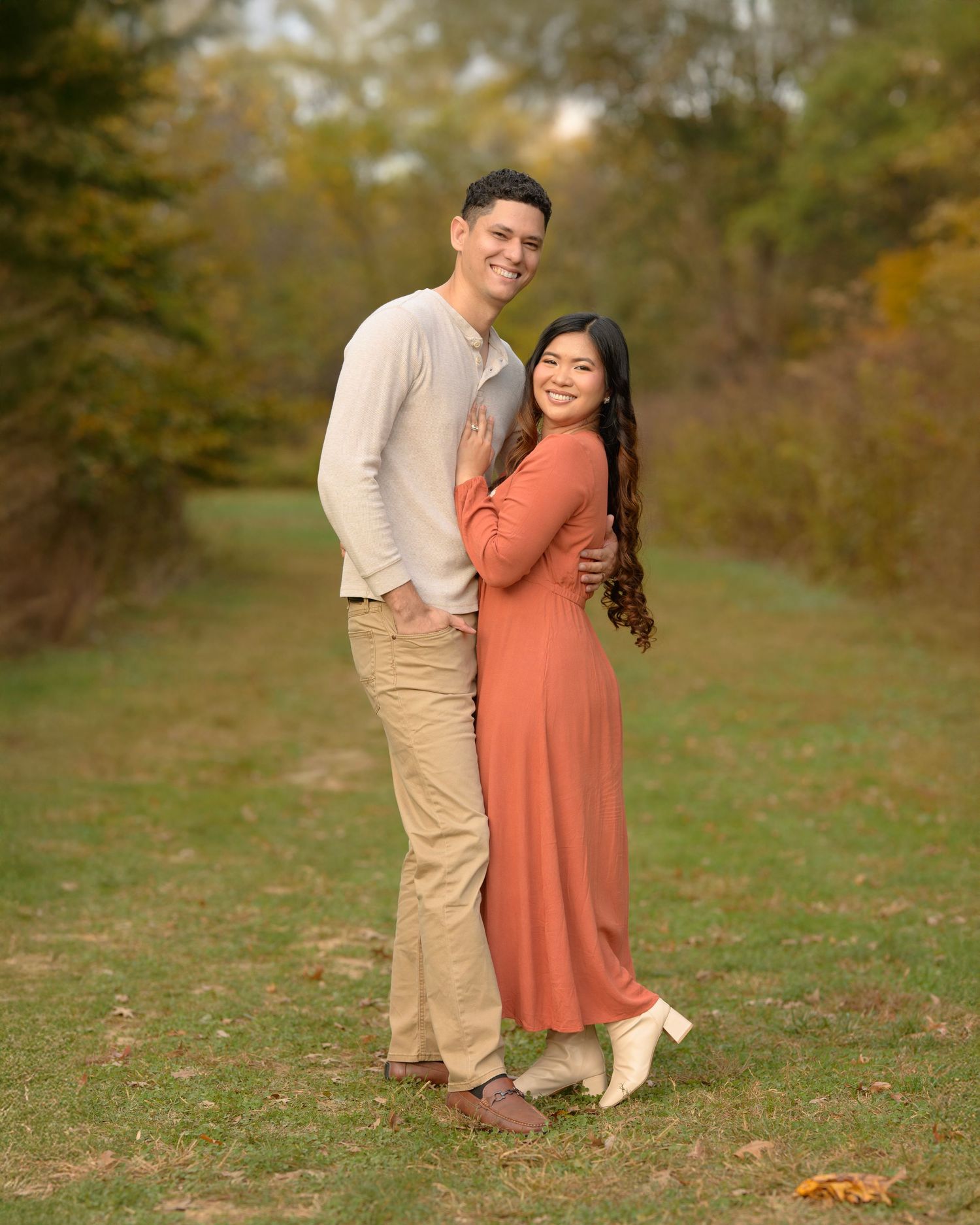 Couple embracing in autumn park, woman in rust dress, man in beige outfit, surrounded by fall foliage.