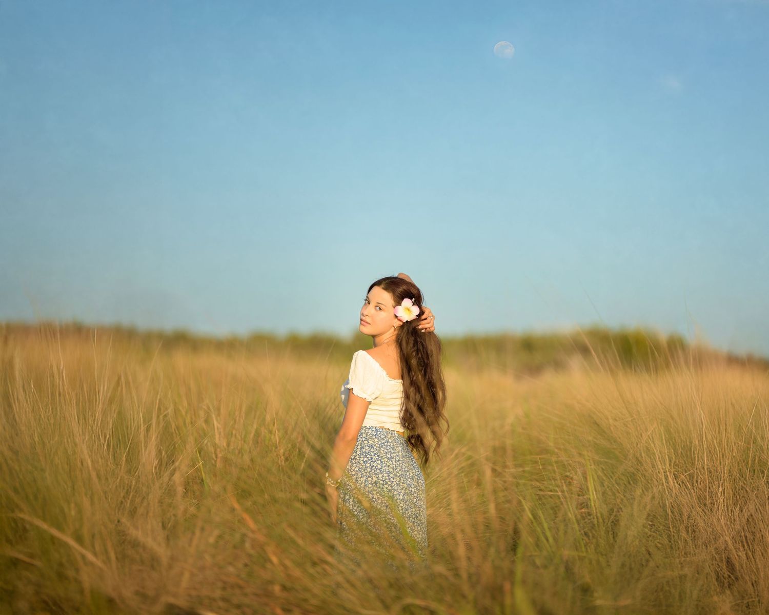 A person in a white dress stands among tall grass in a sunlit field against a blue summer sky.