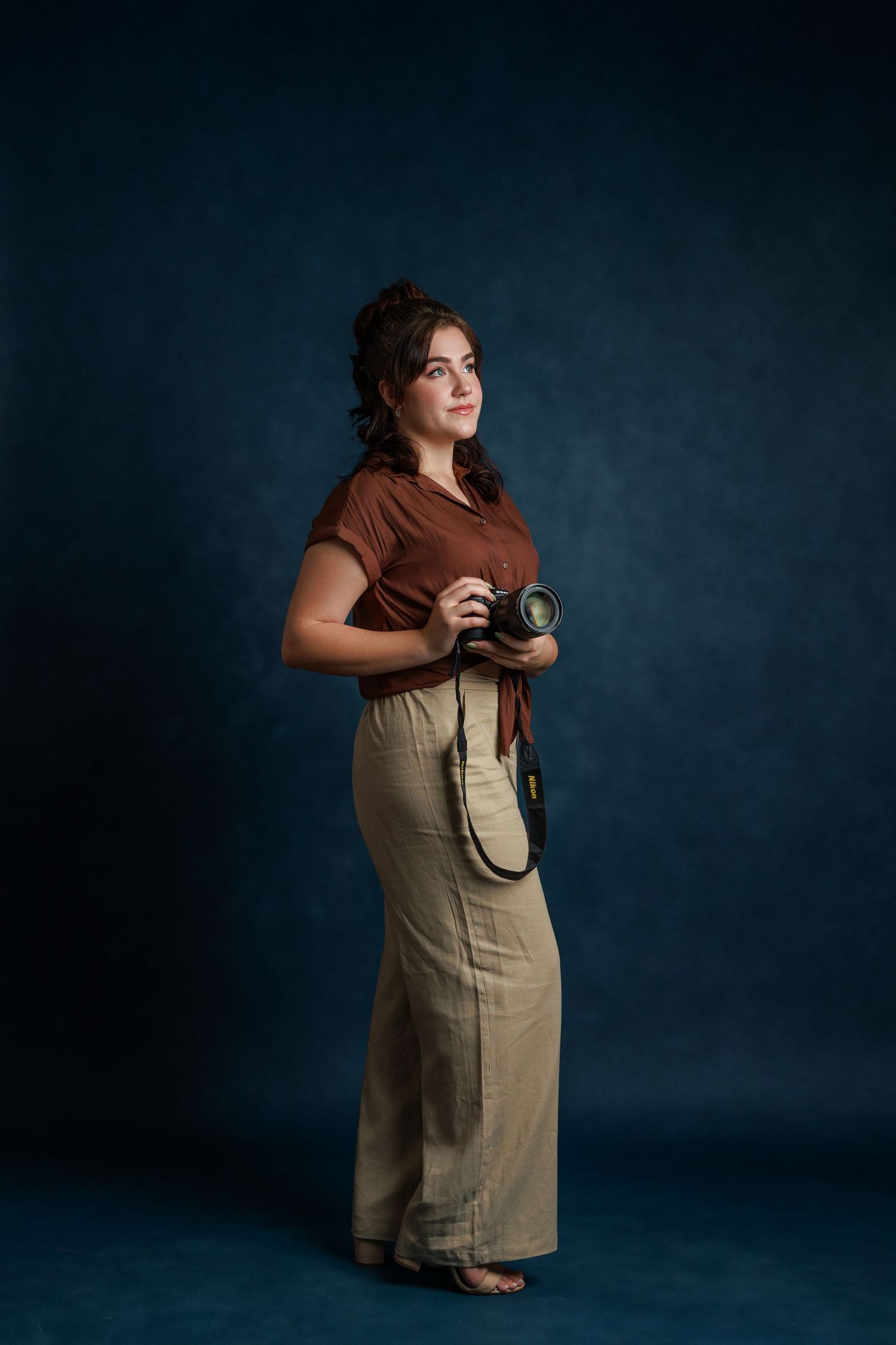 A photographer dressed in a brown shirt and tan overalls poses with a vintage camera against a dark blue background.