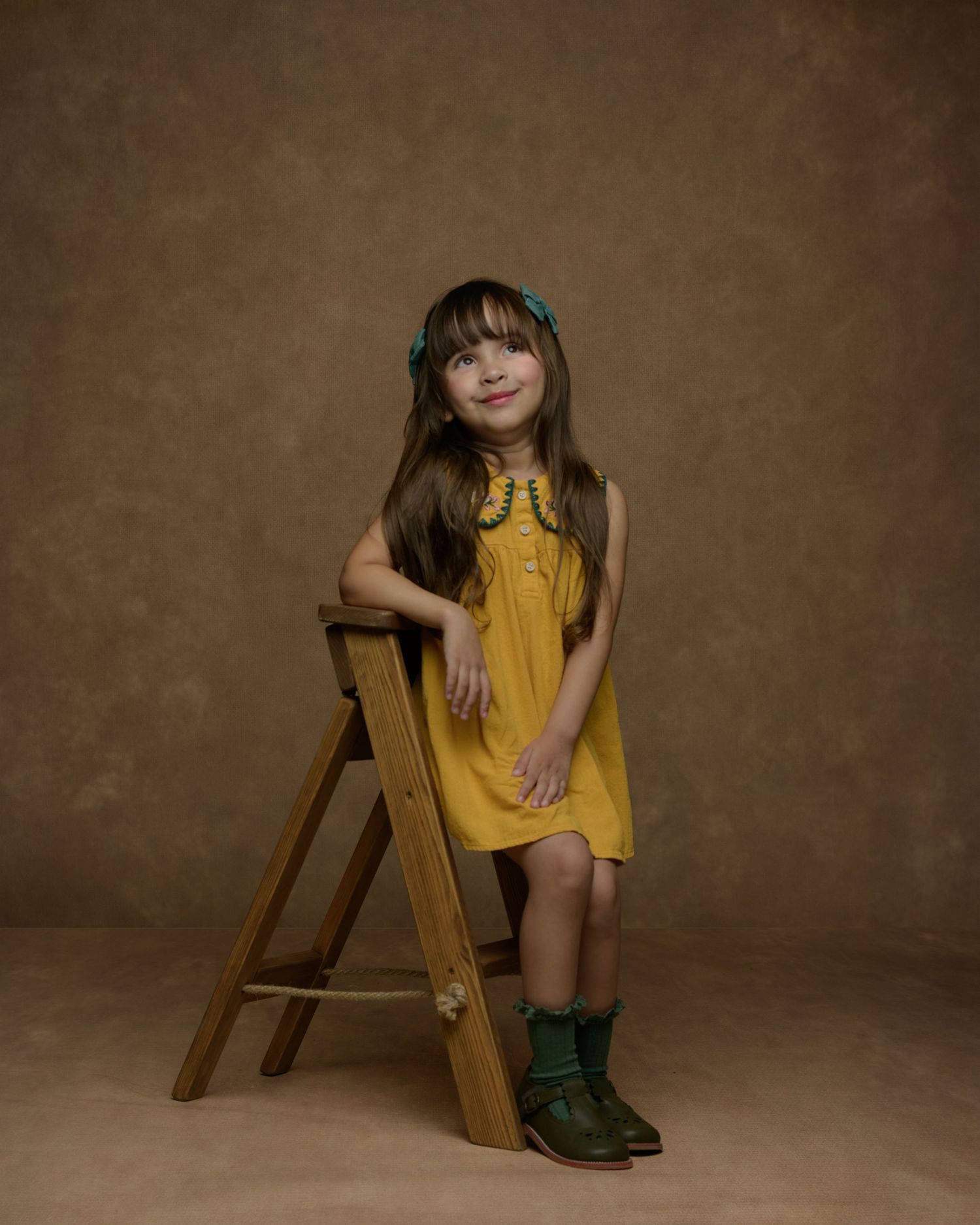 Child in yellow dress sitting on wooden ladder against vintage brown backdrop.