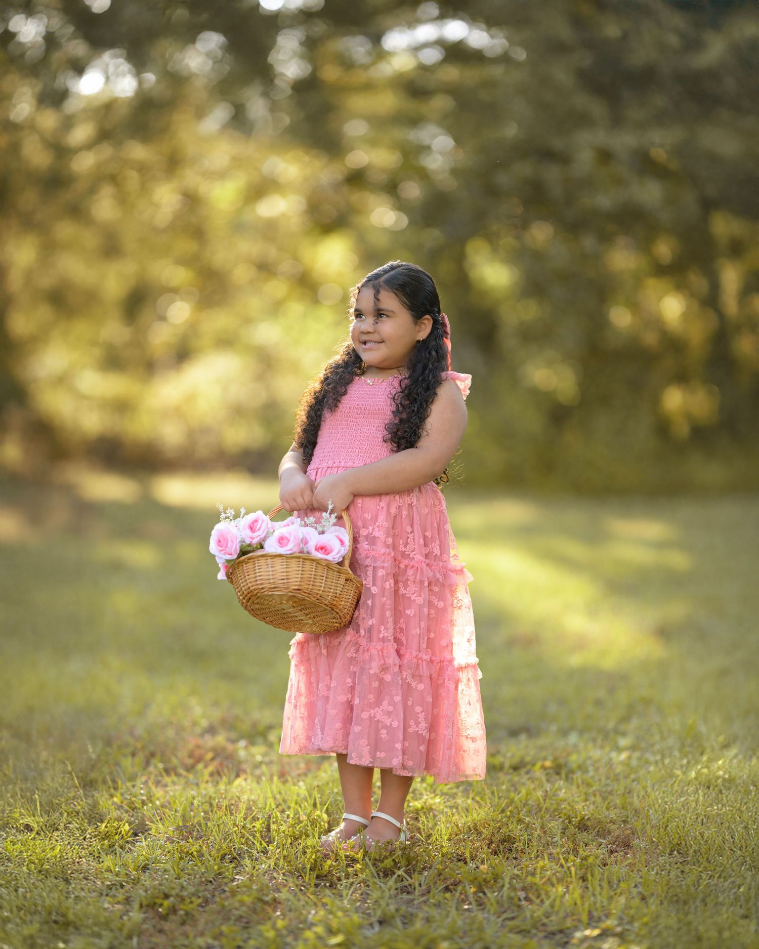 A young girl in a pink dress holds a basket of pink flowers while standing in a sunlit green outdoor setting.