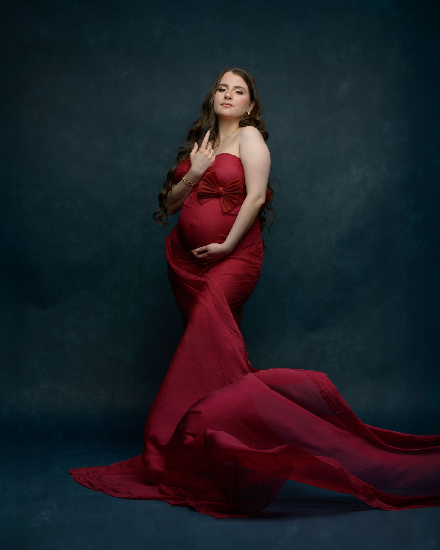 Maternity portrait featuring person in flowing red gown against dark dramatic background.