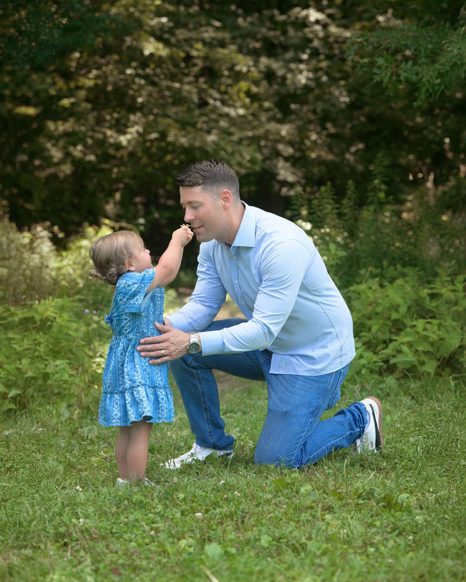 A tender moment captured in nature as a child in a blue dress touches an adult's face while they kneel in green grass.