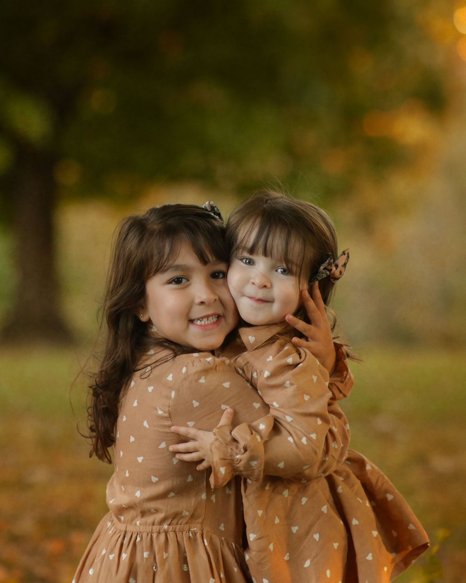Two children in matching brown polka dot dresses embracing outdoors in autumn setting.