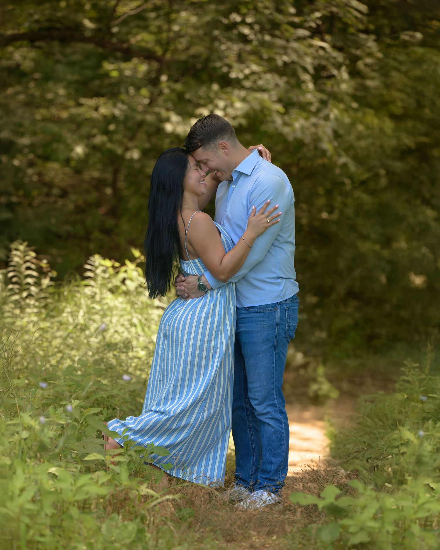 A romantic couple embraces on a sunlit forest path while wearing coordinating blue outfits on a summer afternoon.