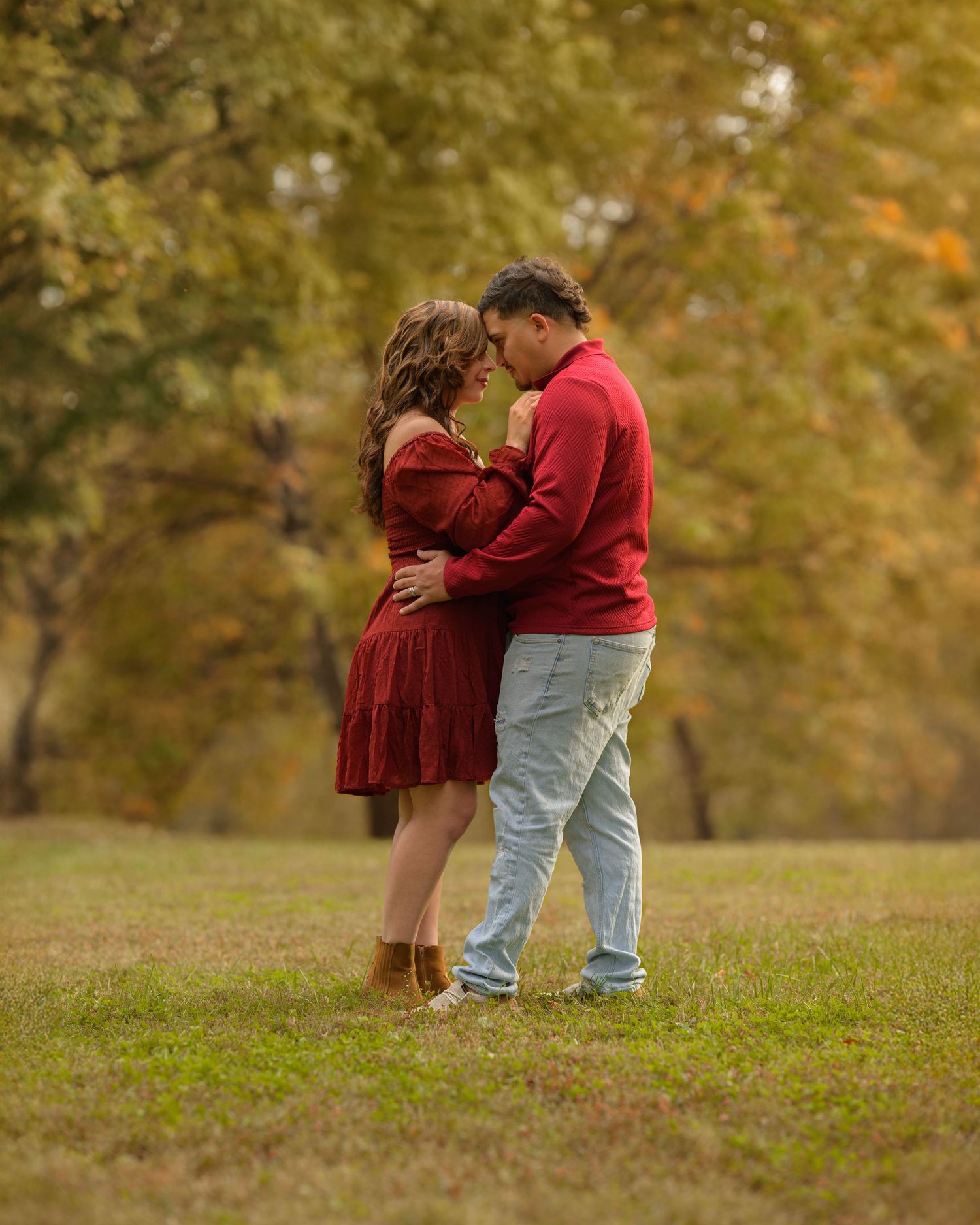 A couple embraces tenderly on a grassy hill surrounded by autumn trees with golden foliage.