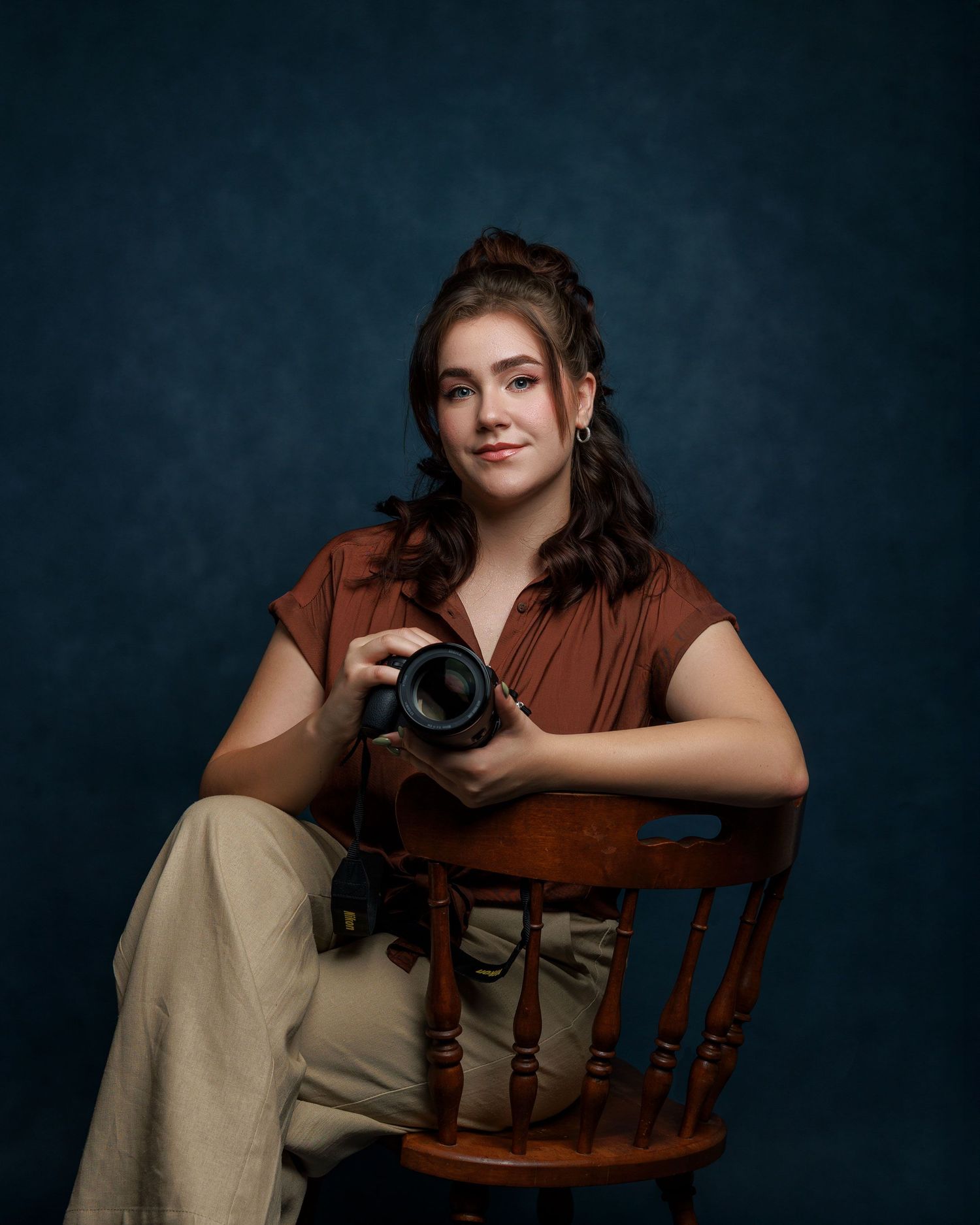 A photographer in a brown jacket and beige top poses with a camera against a dark studio backdrop.