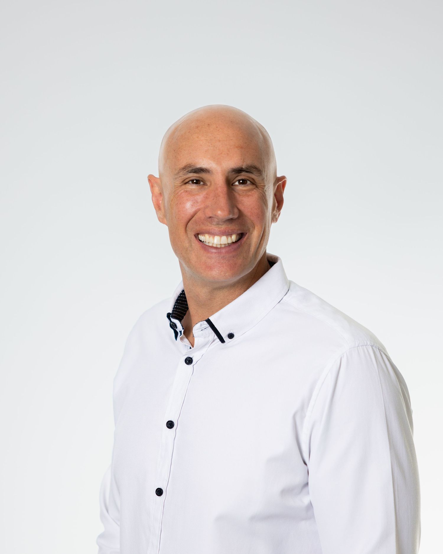 Smiling bald man in a white button-up shirt posing professionally against a light gray background.