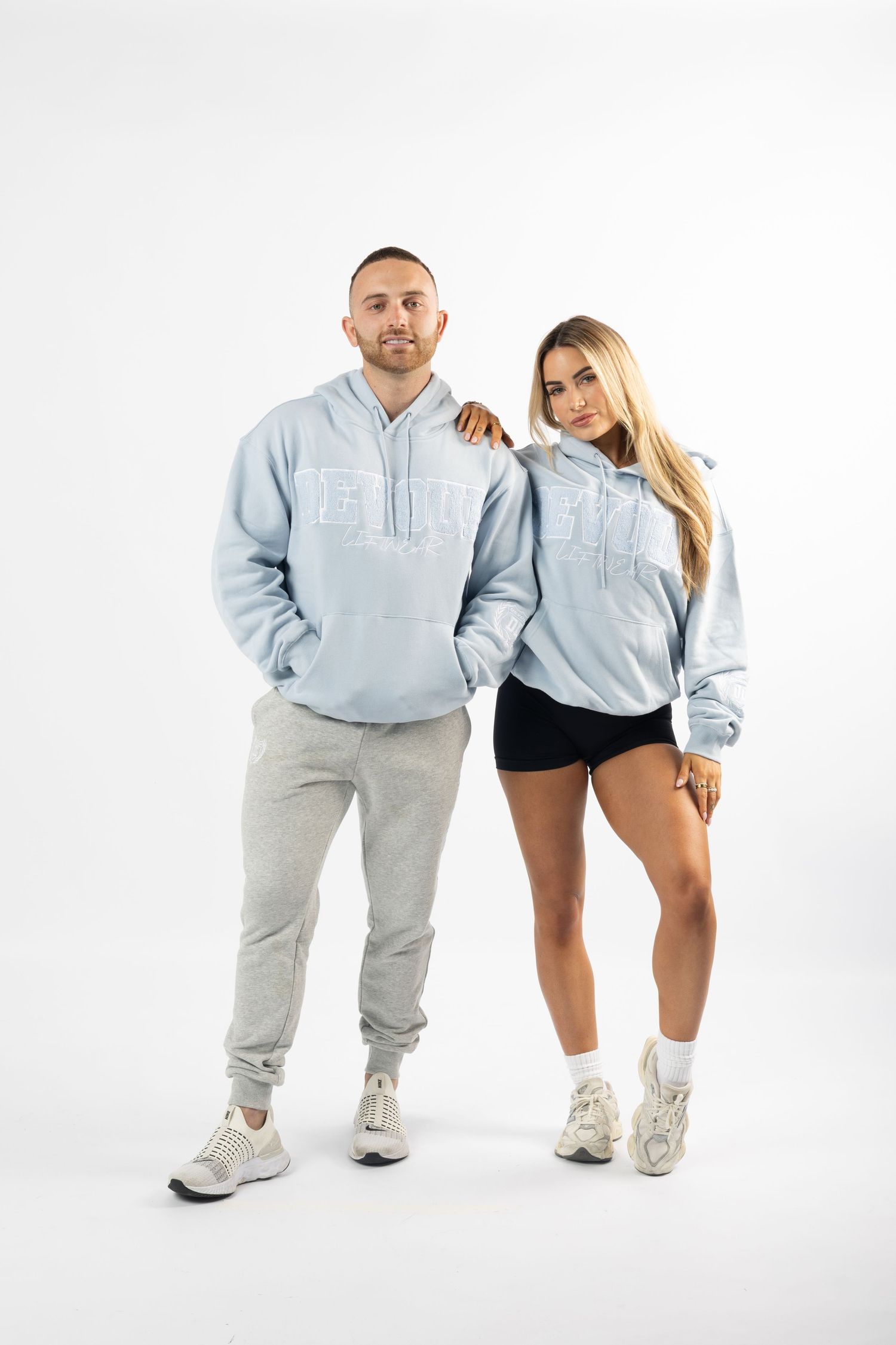 Two people wearing matching light blue hoodies and sneakers pose together against white background.