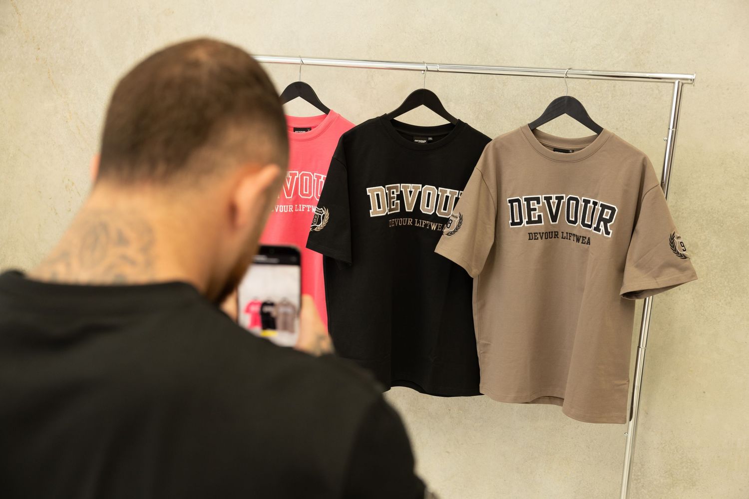 Clothing rack displays three Devour t-shirts in different colors while someone photographs them with a phone.