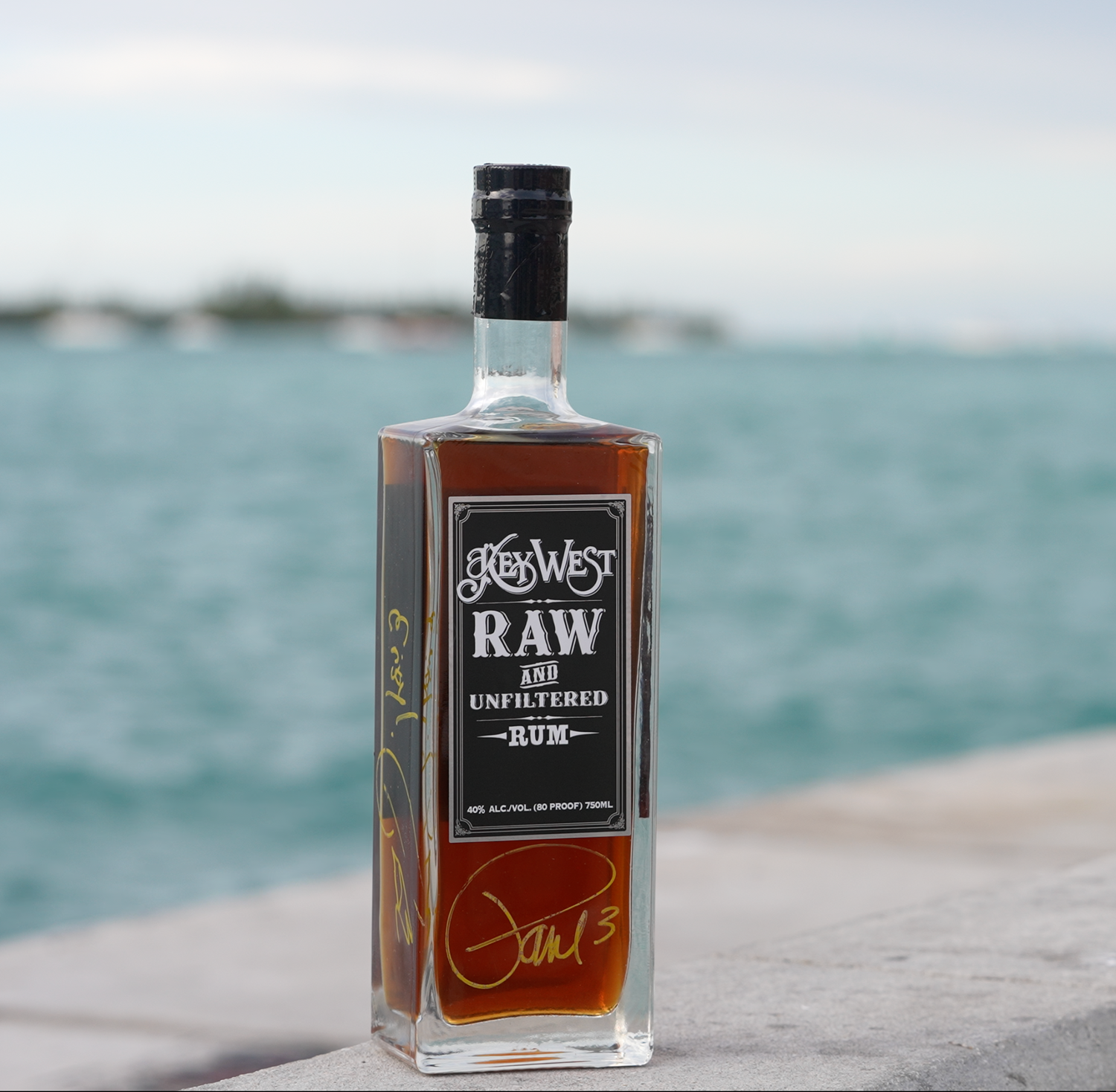 Bottle of JGM's Raw unfiltered rum photographed against blue ocean backdrop.