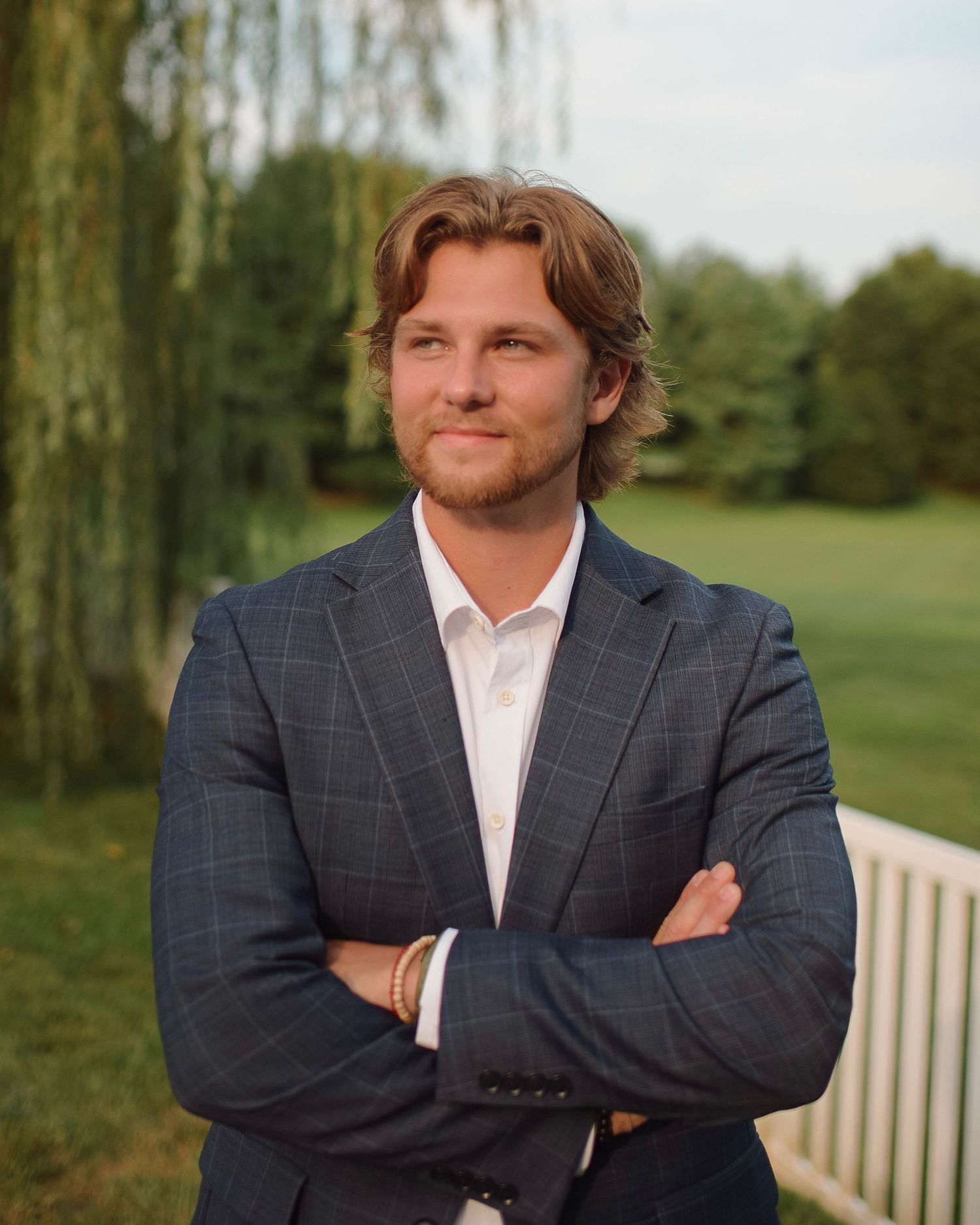 A person in a navy windowpane suit jacket and white shirt stands with arms crossed outside near a willow tree.