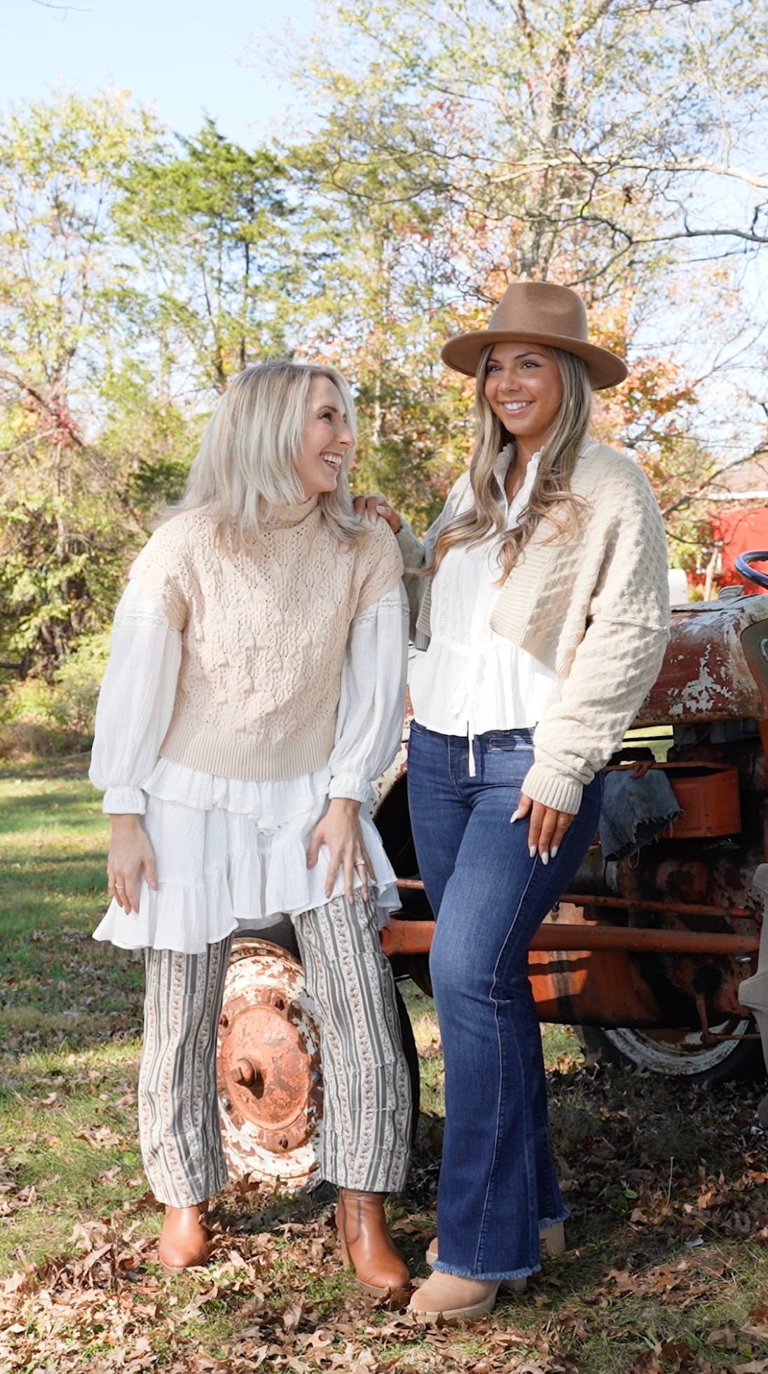 Two women in cozy fall outfits pose by vintage tractor in autumn setting.