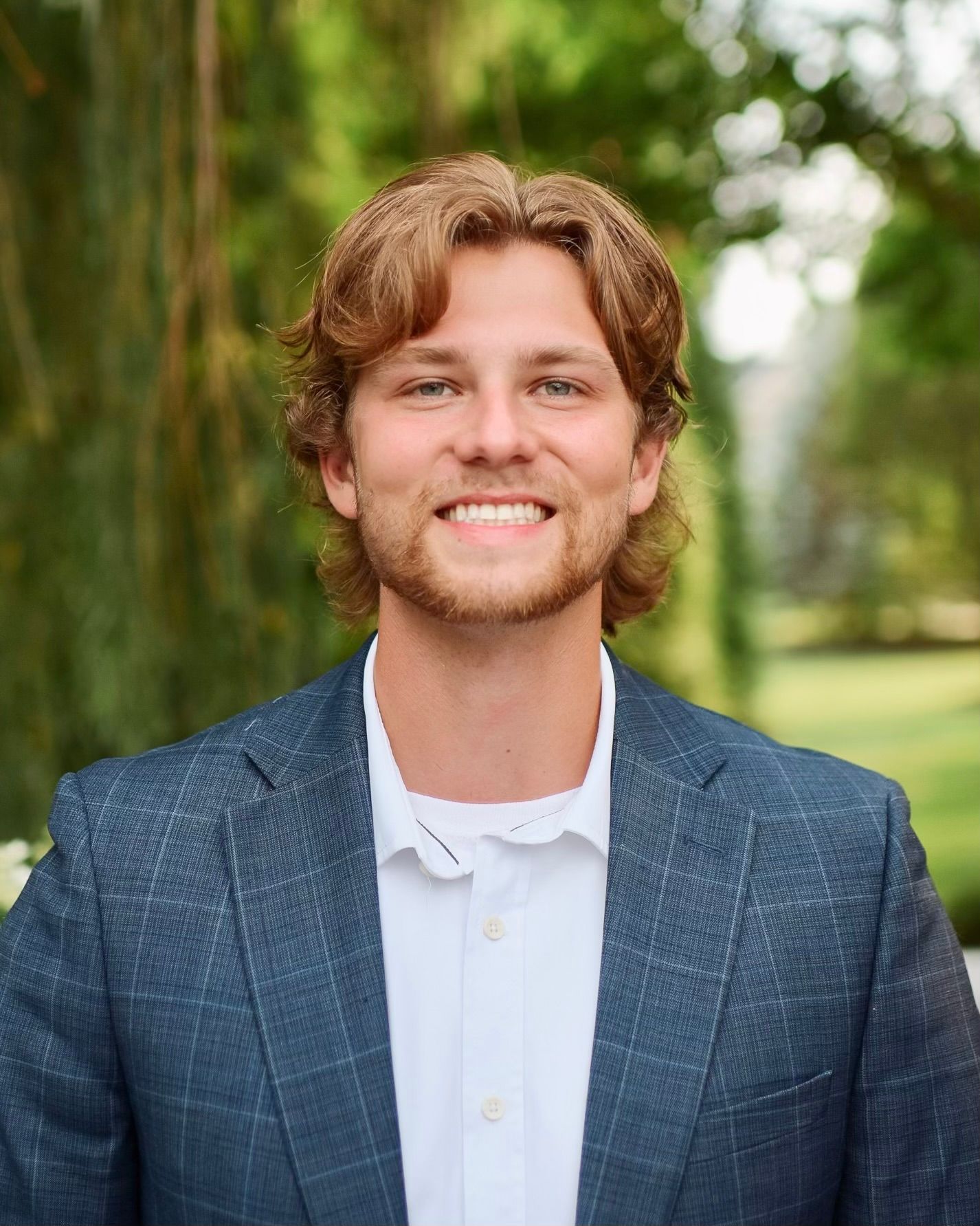 Professional headshot of someone wearing a plaid navy blazer and white shirt against a green outdoor background.