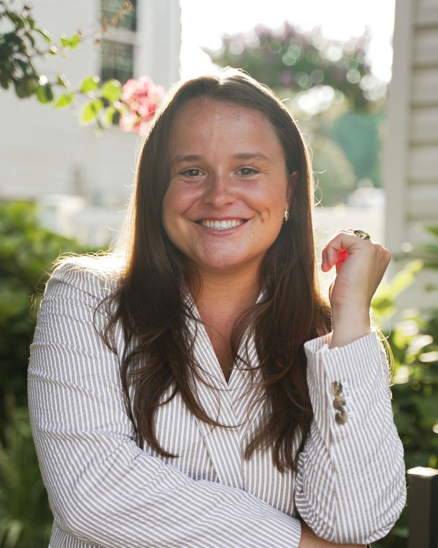 Professional portrait of someone in a white pinstriped button-down shirt sitting outdoors with plants in the background.