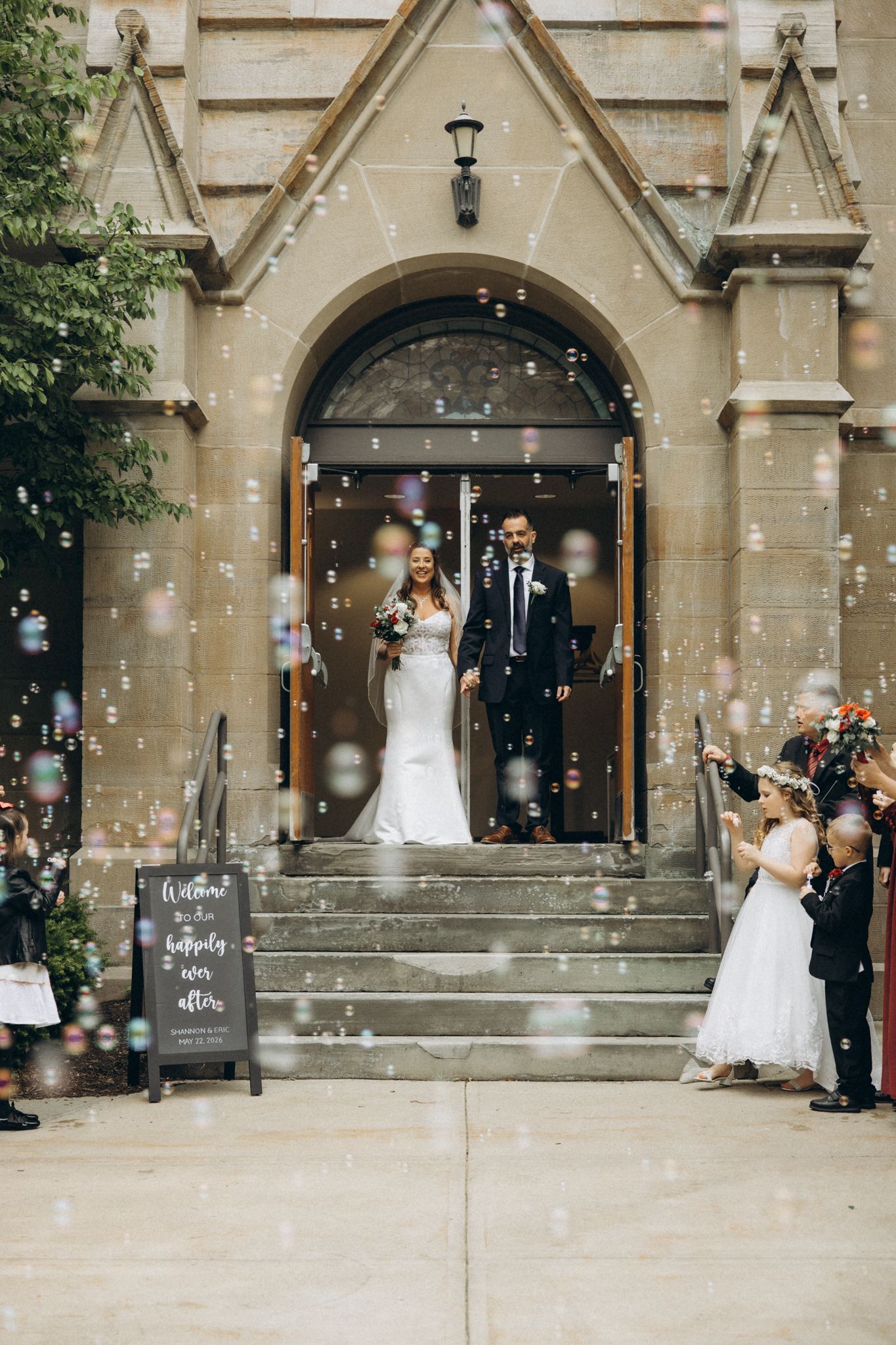 Bride and groom exit stone church surrounded by bubbles as guests celebrate their wedding ceremony.
