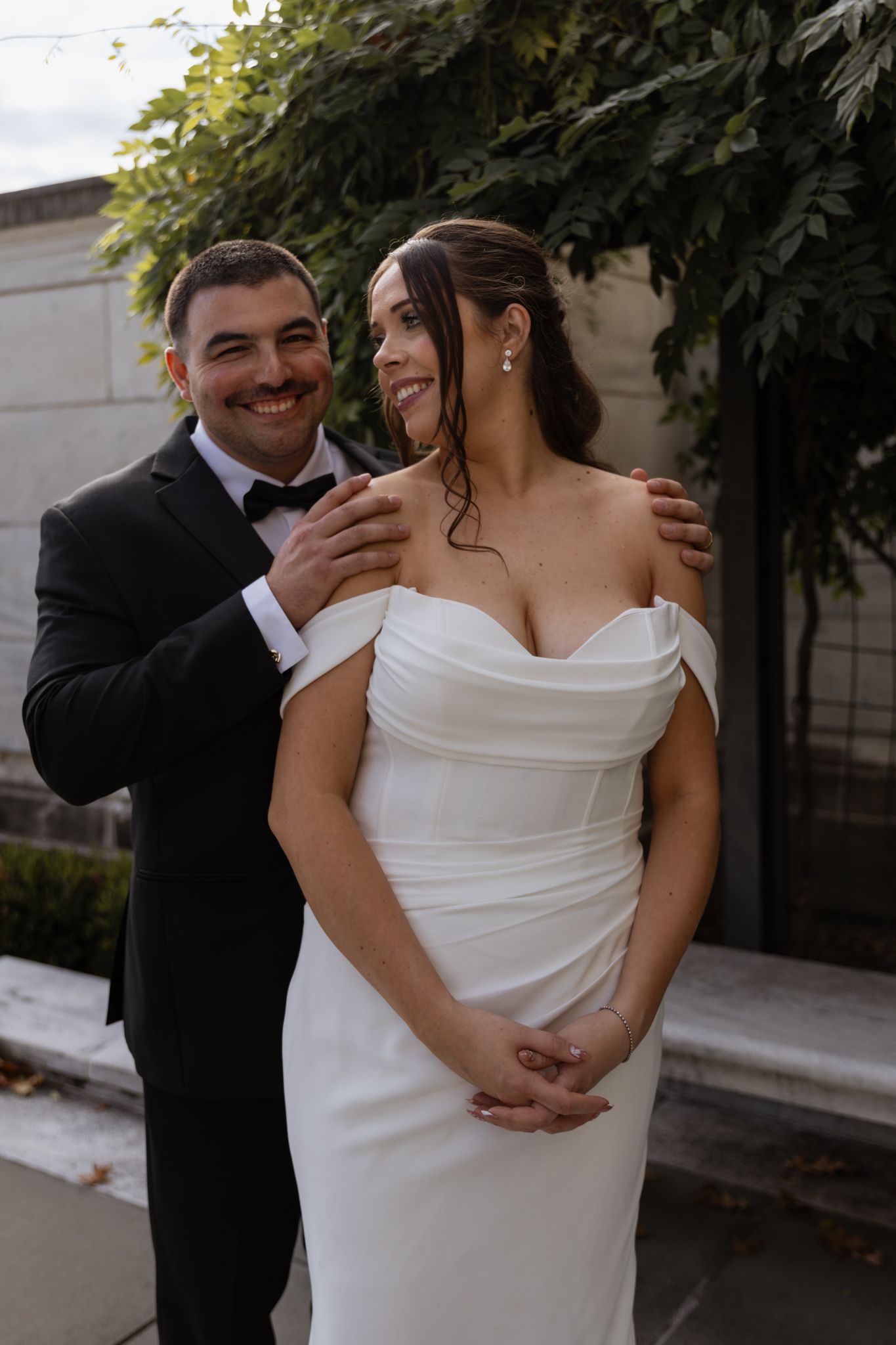 Bride in off-shoulder white gown and groom in black tuxedo smiling together outdoors at their wedding.