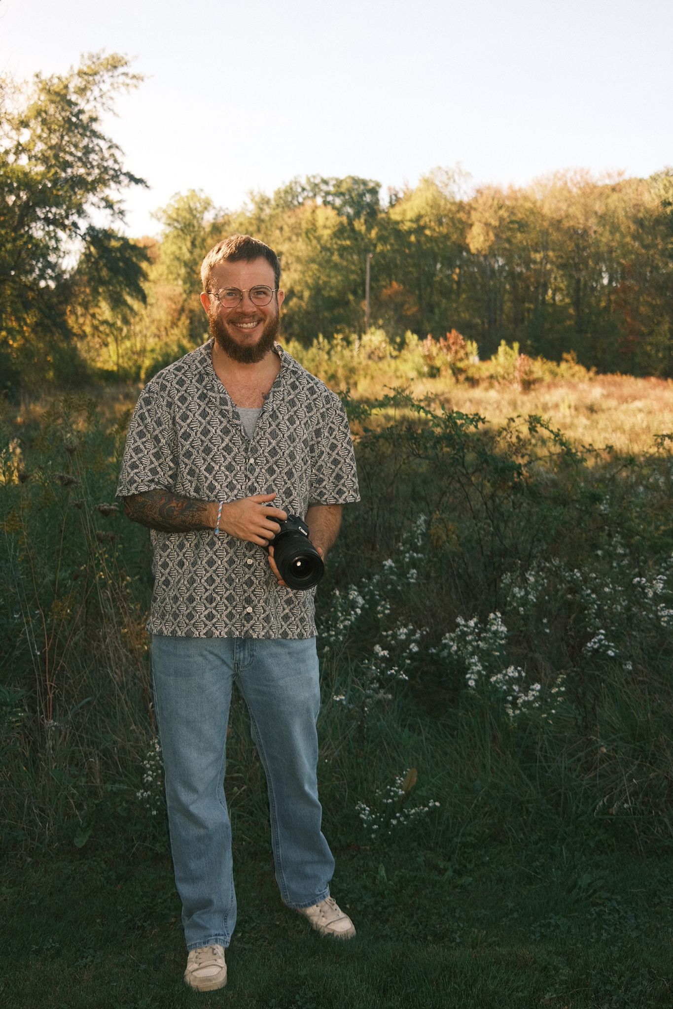 A photographer in patterned shirt and jeans holds camera in outdoor setting during golden hour.