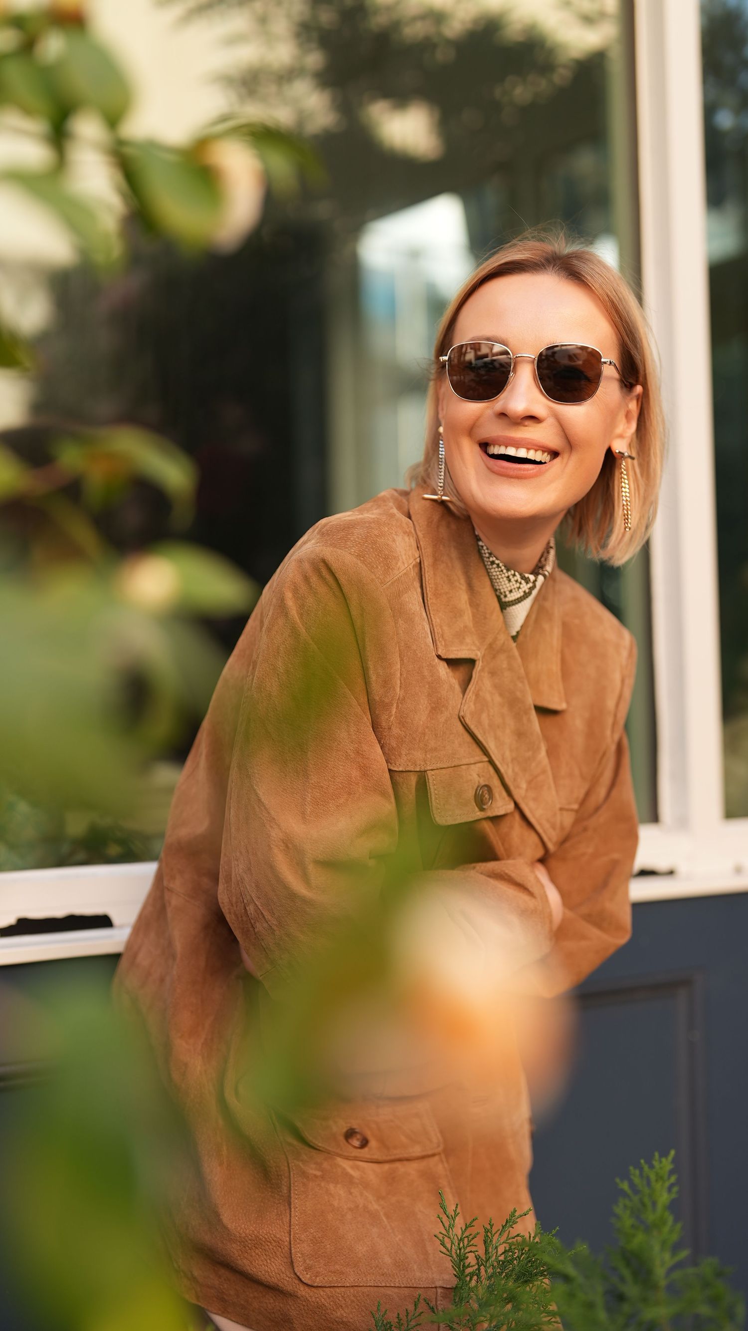 Woman in tan suede jacket and sunglasses smiling outdoors among greenery.