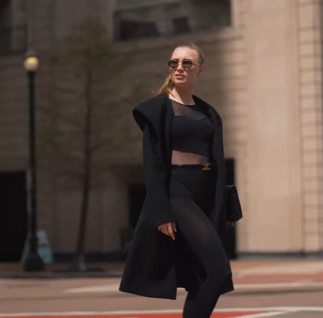 Street style fashion photo of model in black crop top and long coat against urban backdrop.