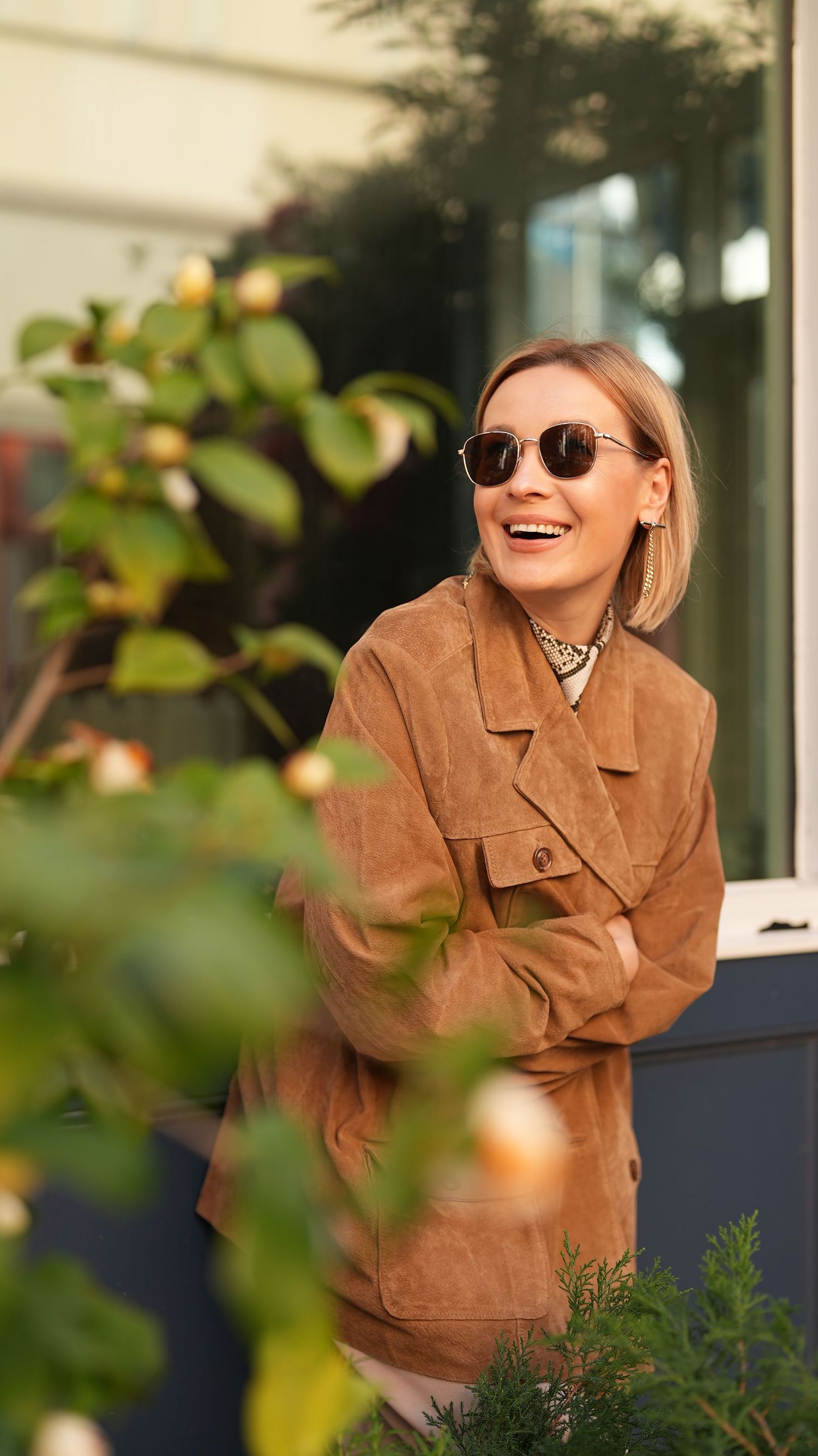 Stylish woman in brown suede jacket and sunglasses laughing outdoors by green foliage.