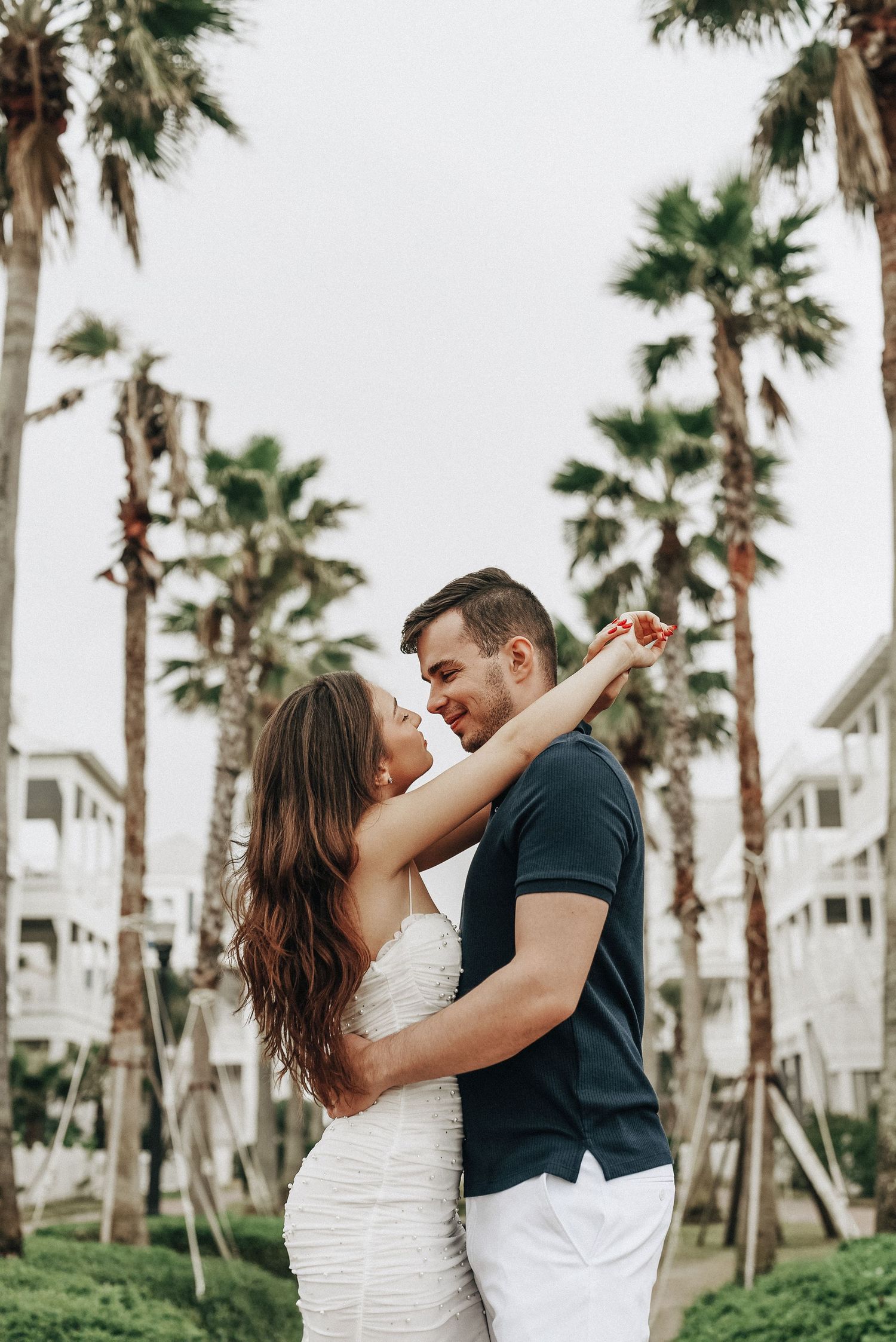 A romantic embrace under tall palm trees with white buildings in the background.