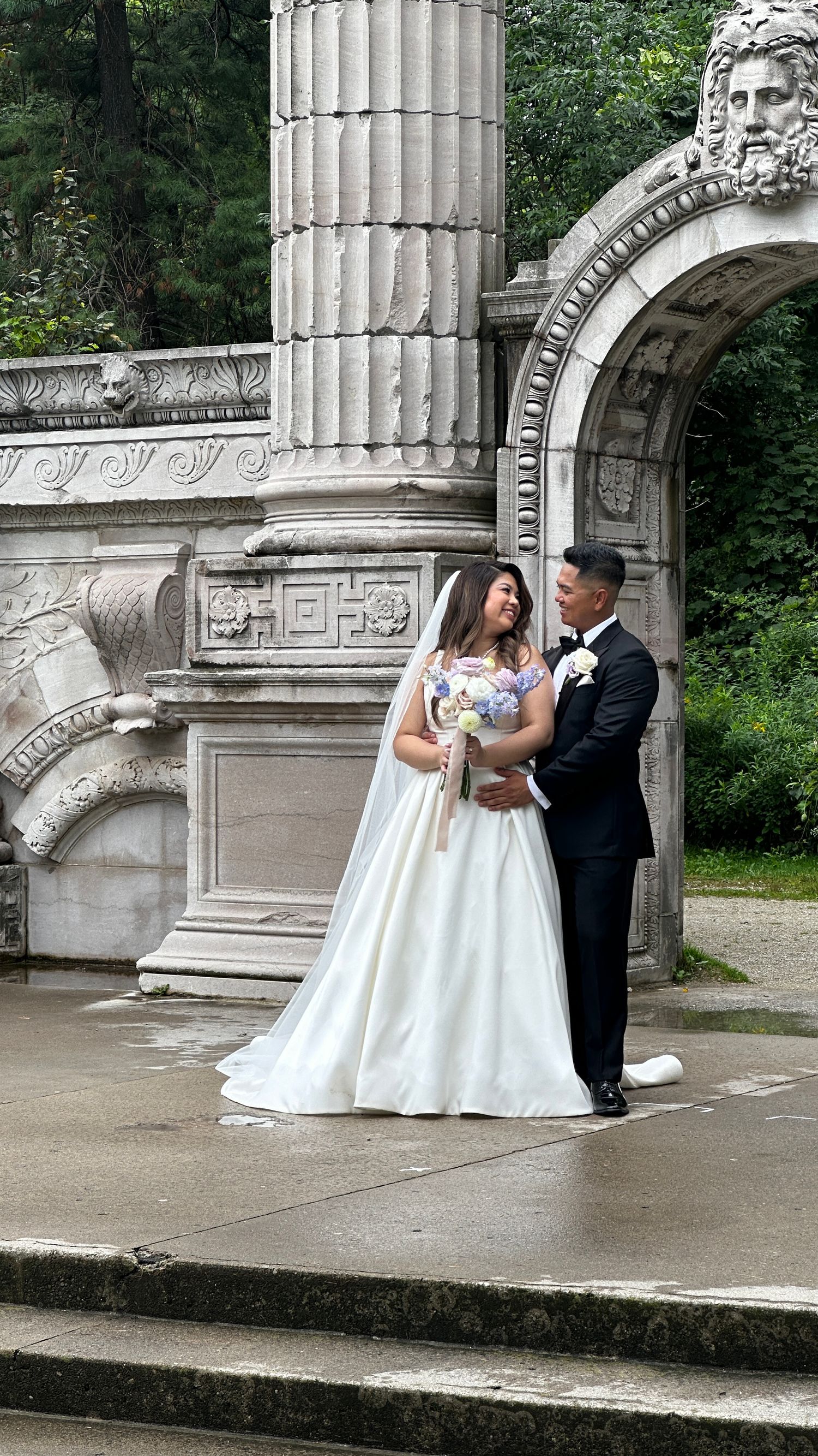 Wedding couple poses beneath ornate classical stone archway and columns in garden setting.