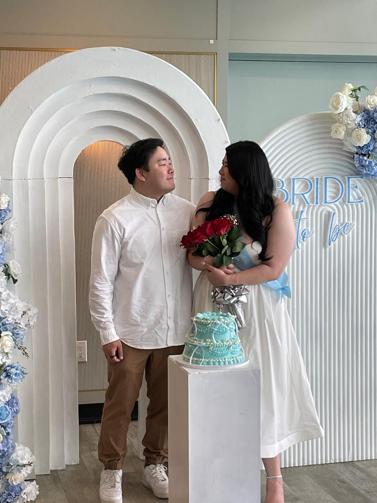 A couple poses together in front of a white arch with blue floral decorations and a turquoise cake display.