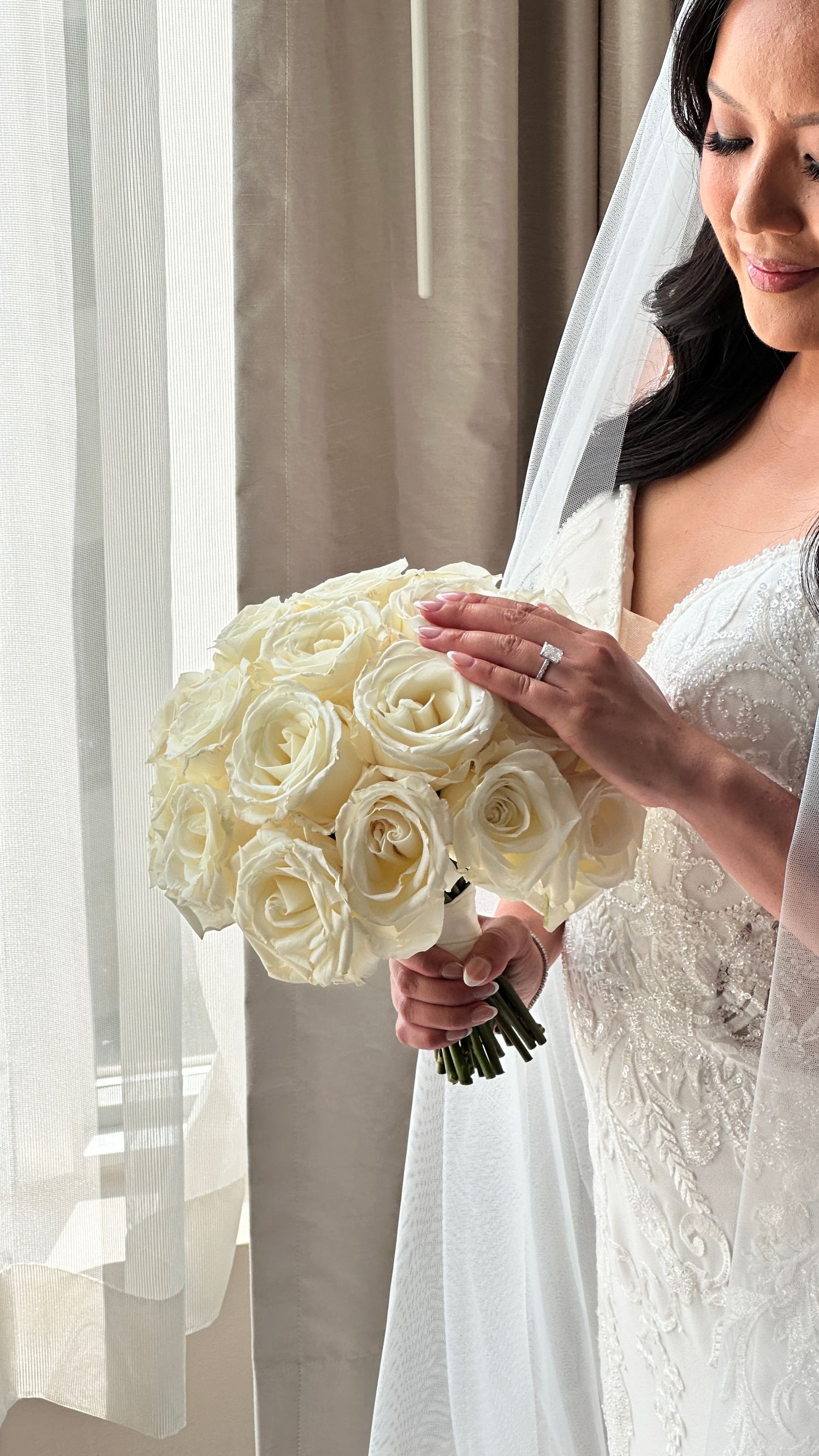 A bride in a white lace dress holds an elegant bouquet of cream-colored roses near a window with sheer curtains.