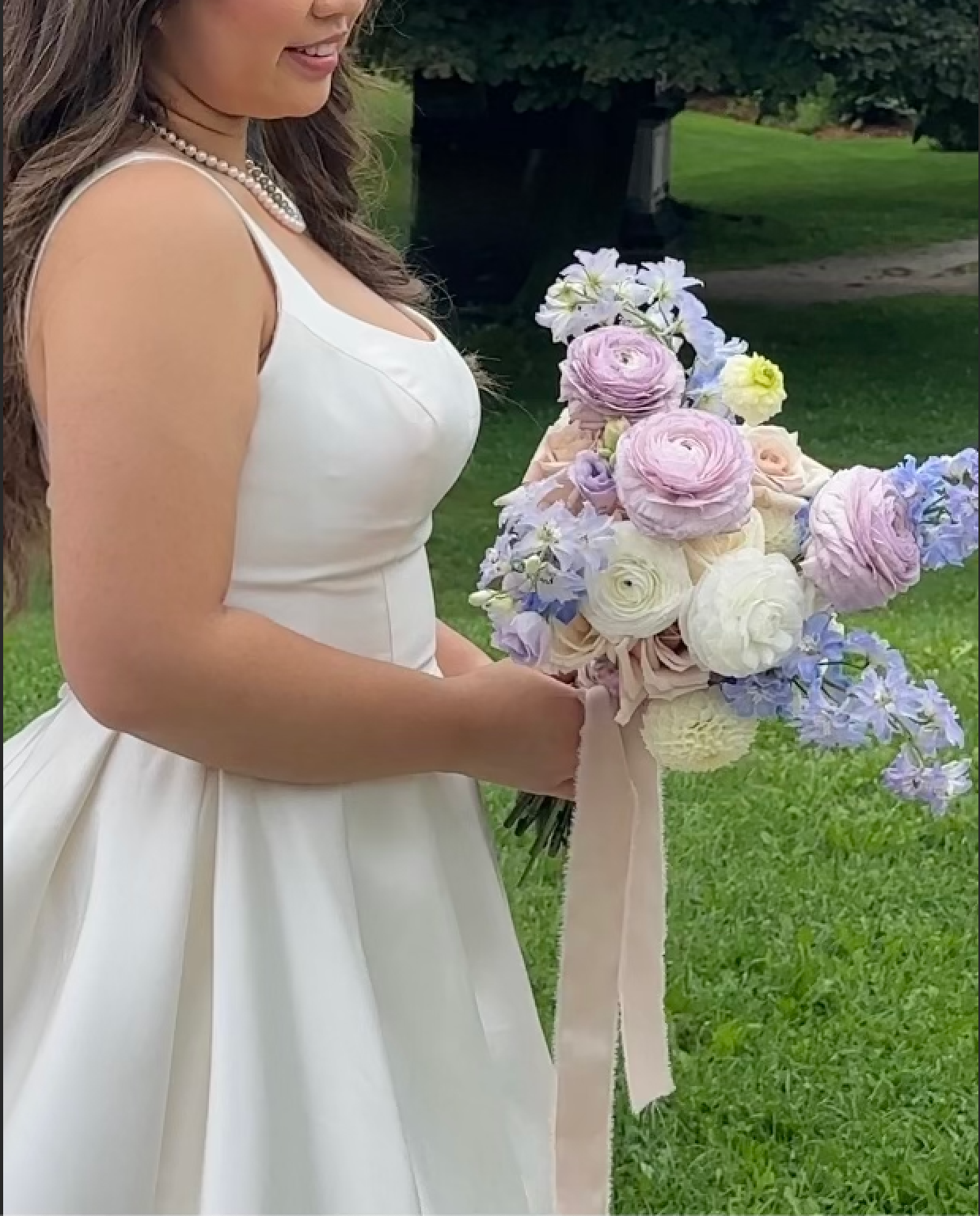 A bridal bouquet featuring soft purple and white flowers with trailing ribbons held against a white dress outdoors.