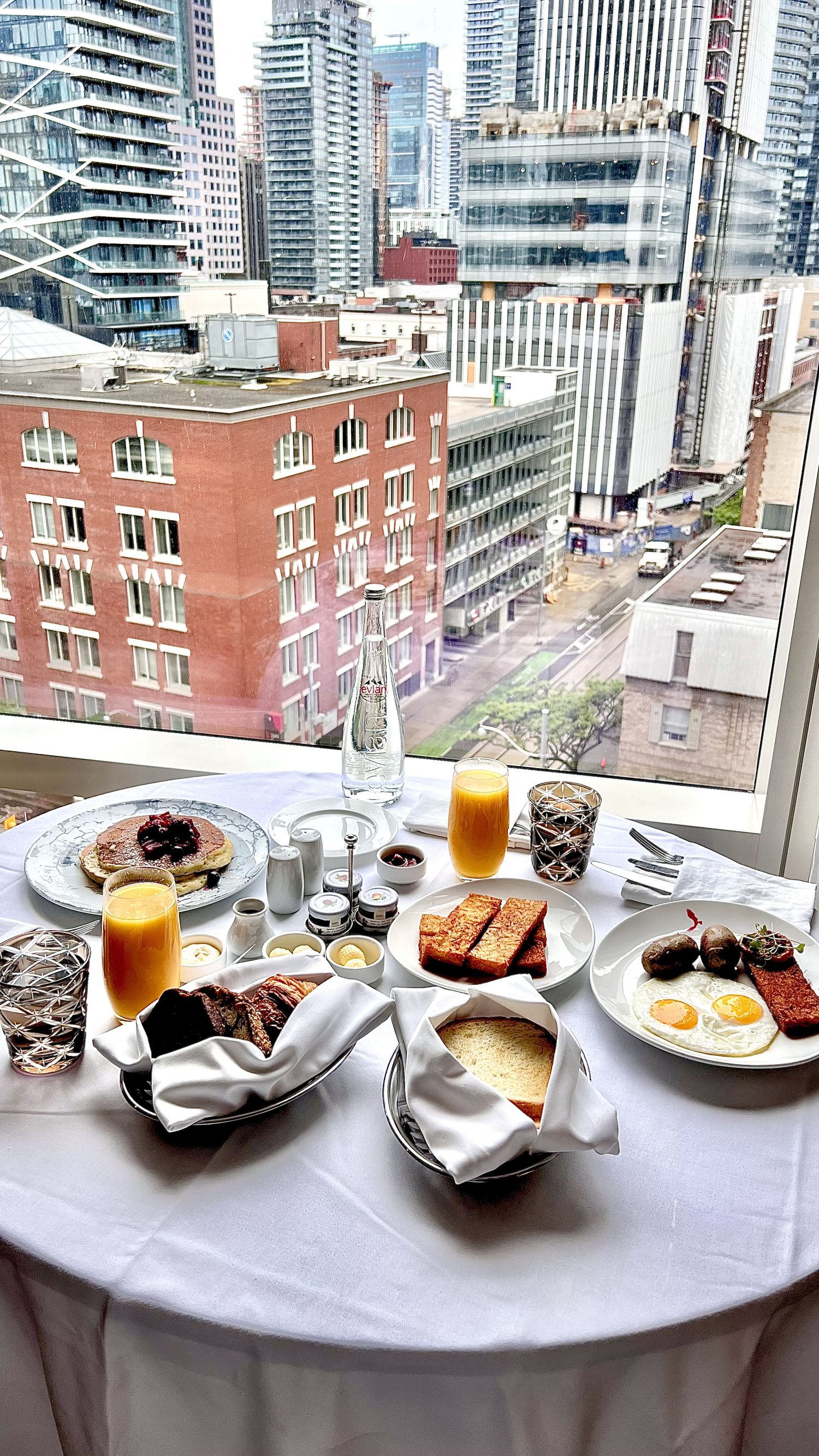 Luxurious breakfast spread with eggs, pancakes and orange juice on a white tablecloth overlooking urban high-rise buildings.