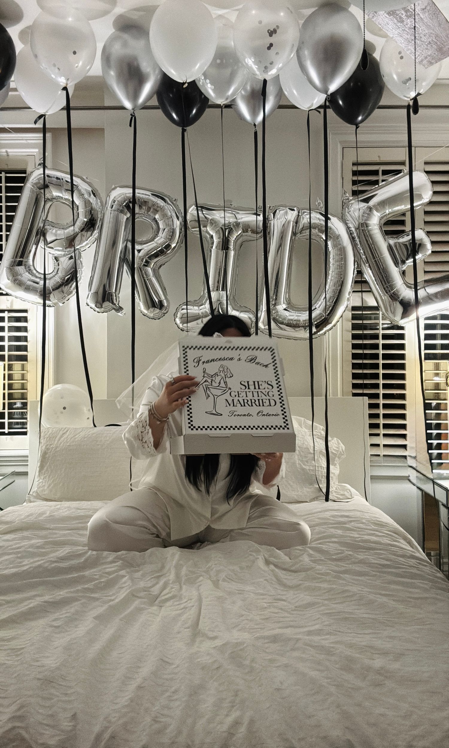 Silver 'BRIDE' balloon letters float above a bed with black and white balloons in a bridal celebration setup.