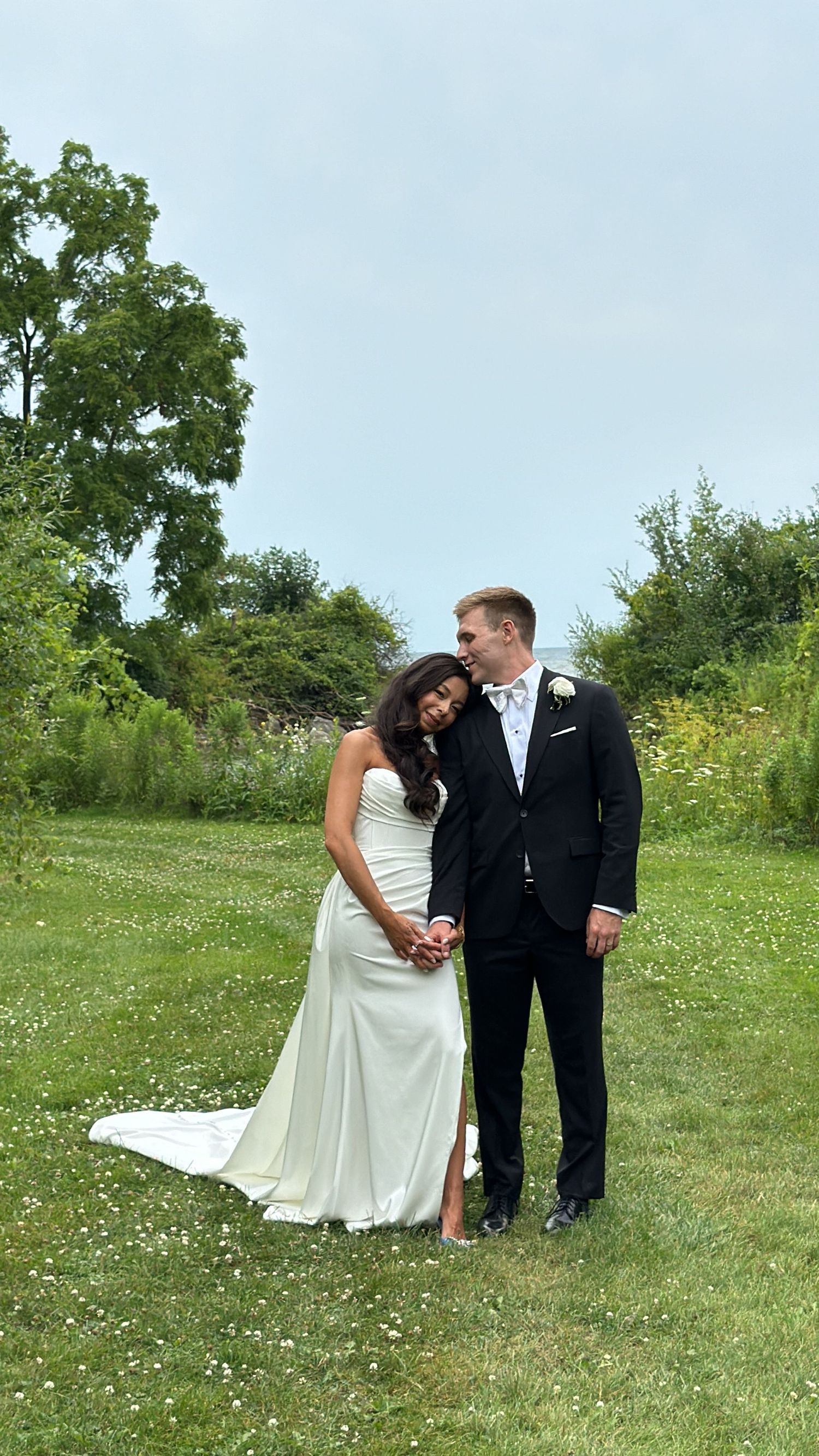 Couple shares intimate wedding moment in natural meadow setting with trees in background.