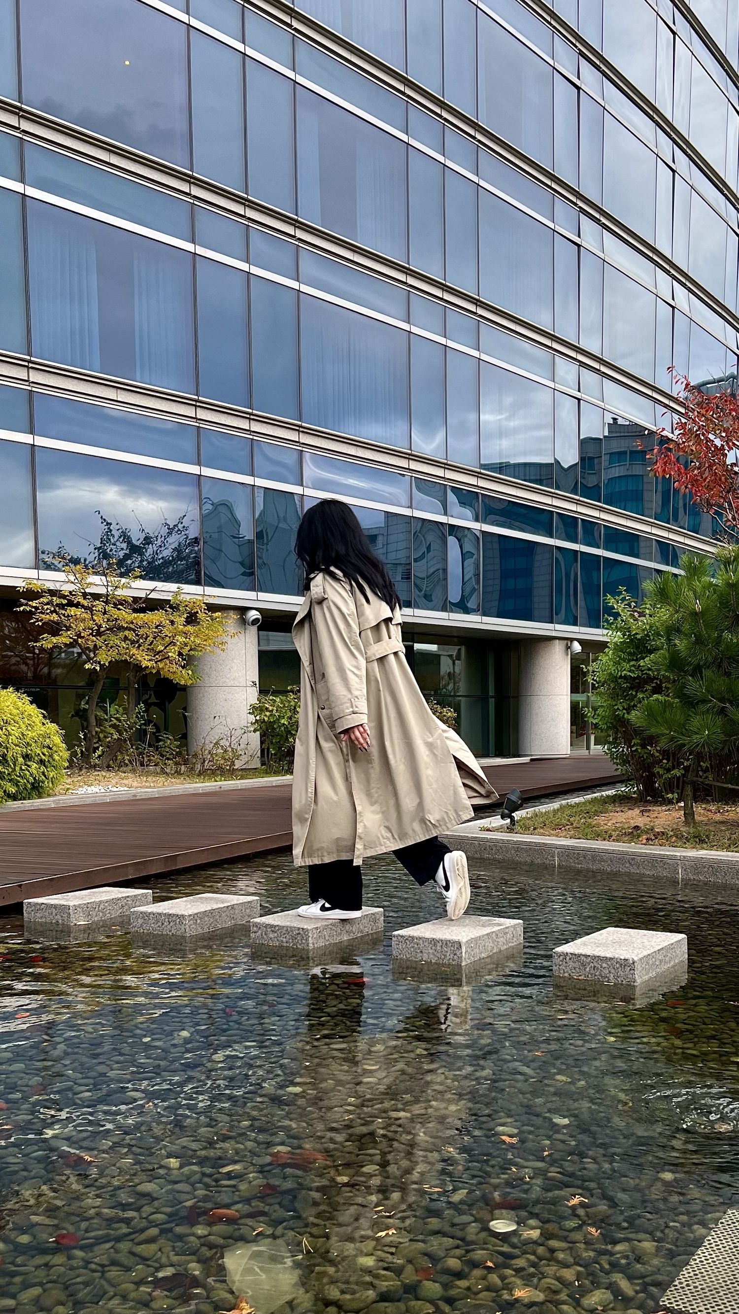 A series of architectural columns along a modern glass building reflected in a shallow decorative pond with stones.