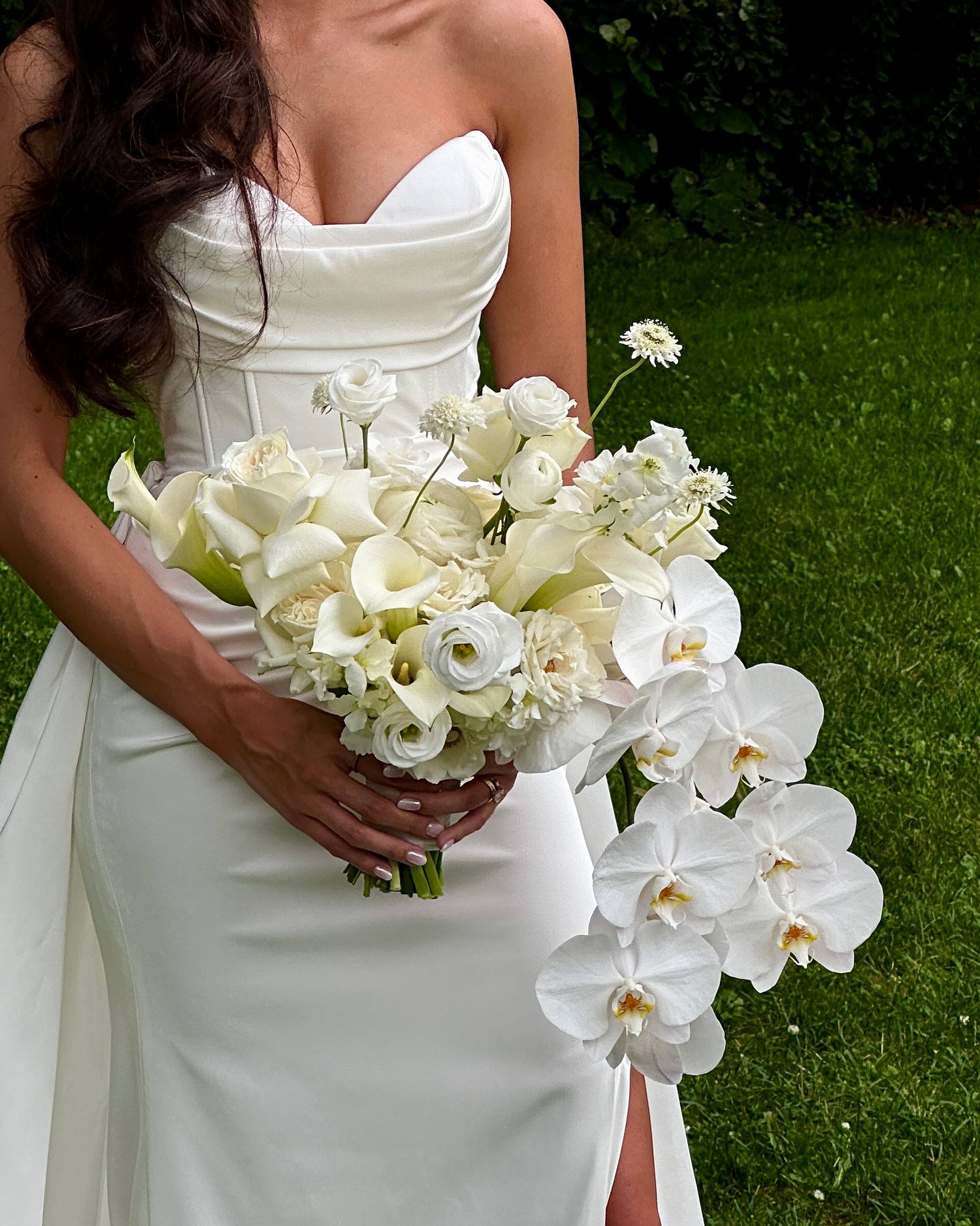Elegant white orchid and calla lily bridal bouquet held against a white wedding dress with green lawn background.