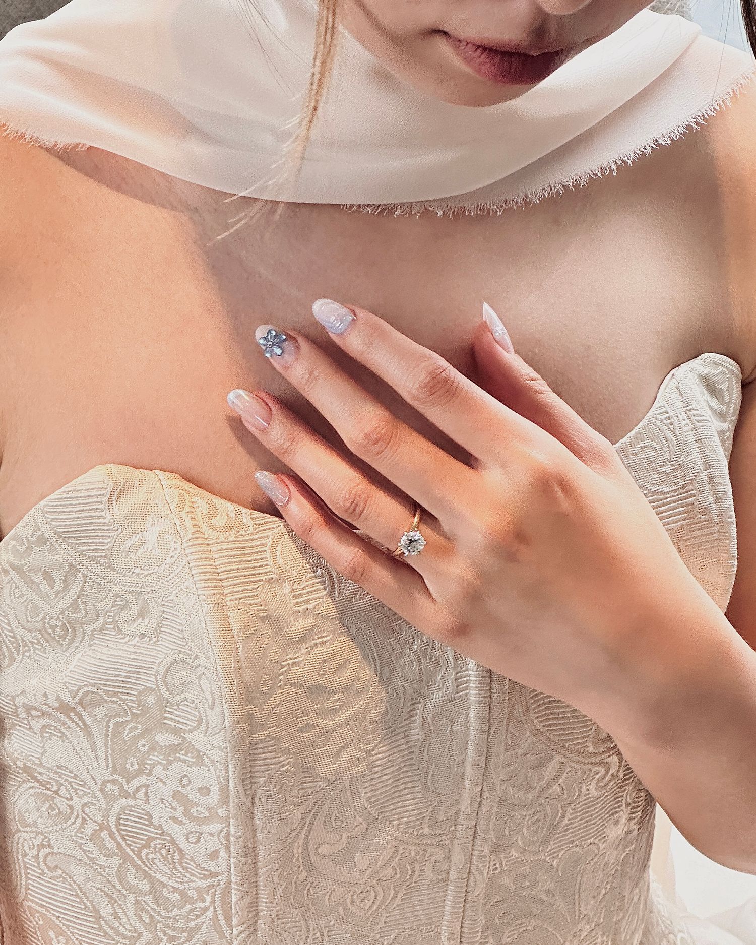 Close-up of a hand with an engagement ring and manicured nails against a white lace dress.