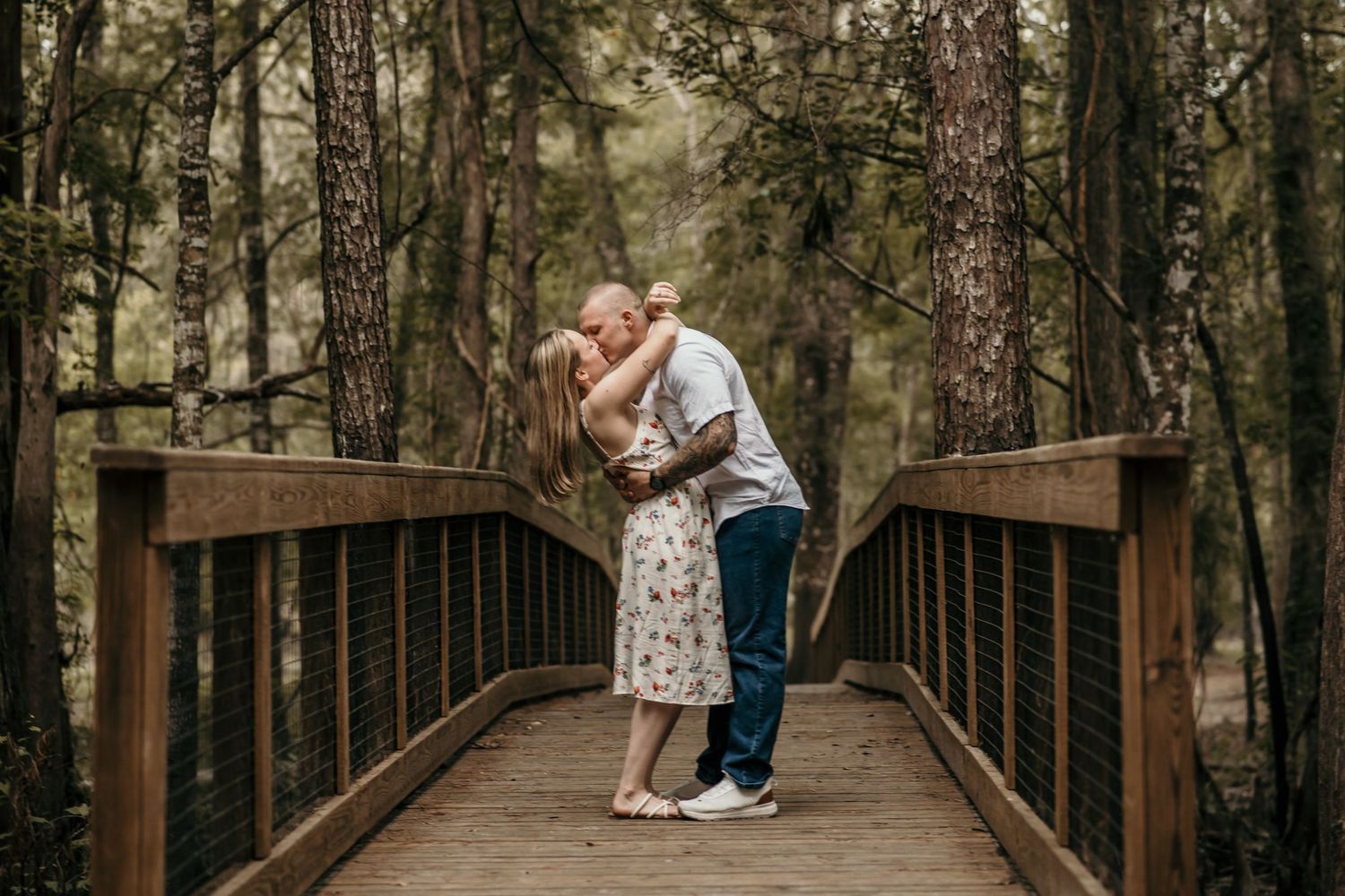 Couple embracing on wooden bridge surrounded by tall forest trees.