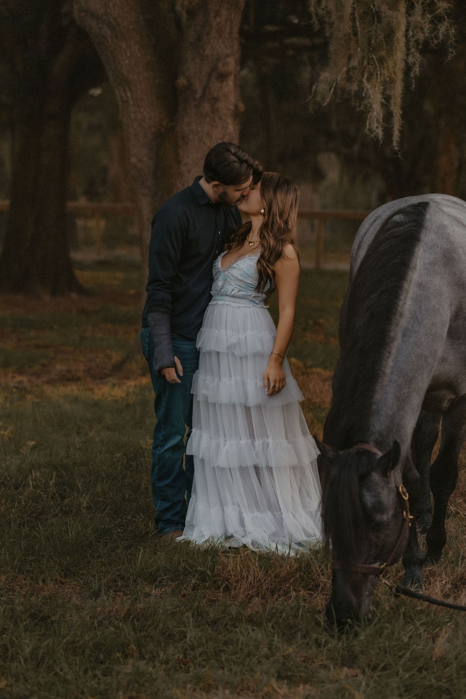 A couple in formal attire stands near a horse in a moody forest setting during sunset.