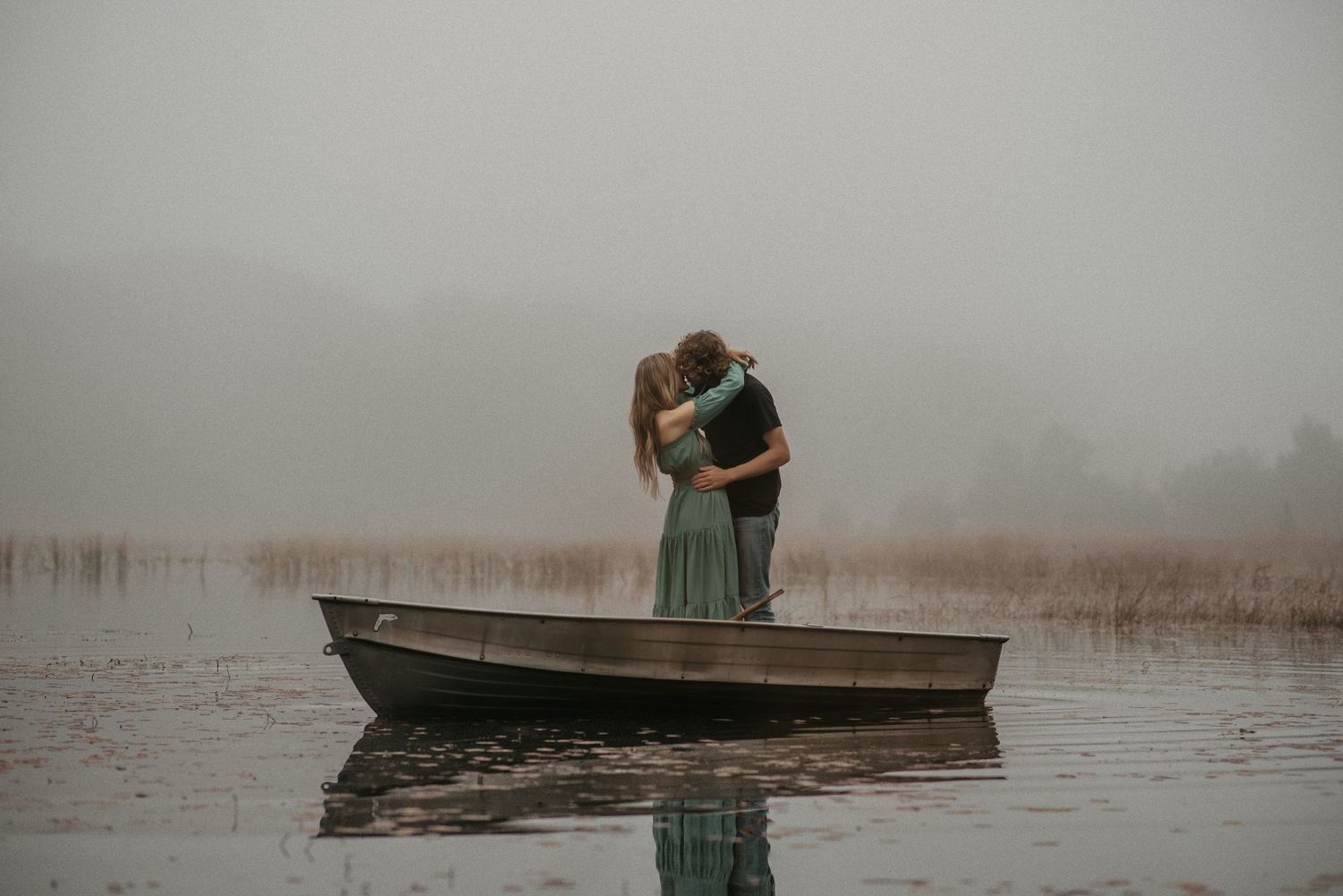 Romantic couple embraces in a wooden boat on misty lake during foggy morning.