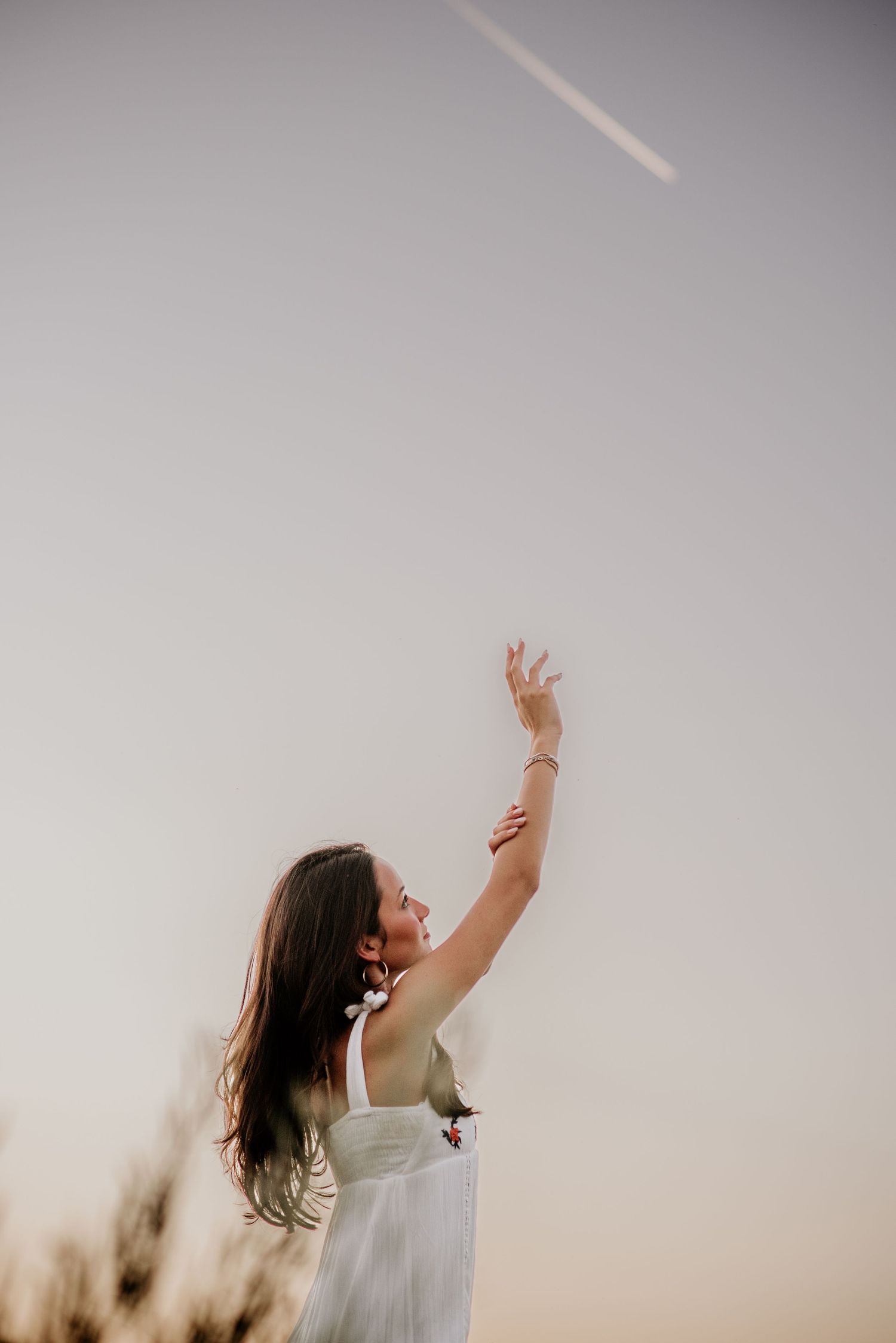 A woman in a white dress reaches upward with graceful movement against a soft, dreamy sky background.