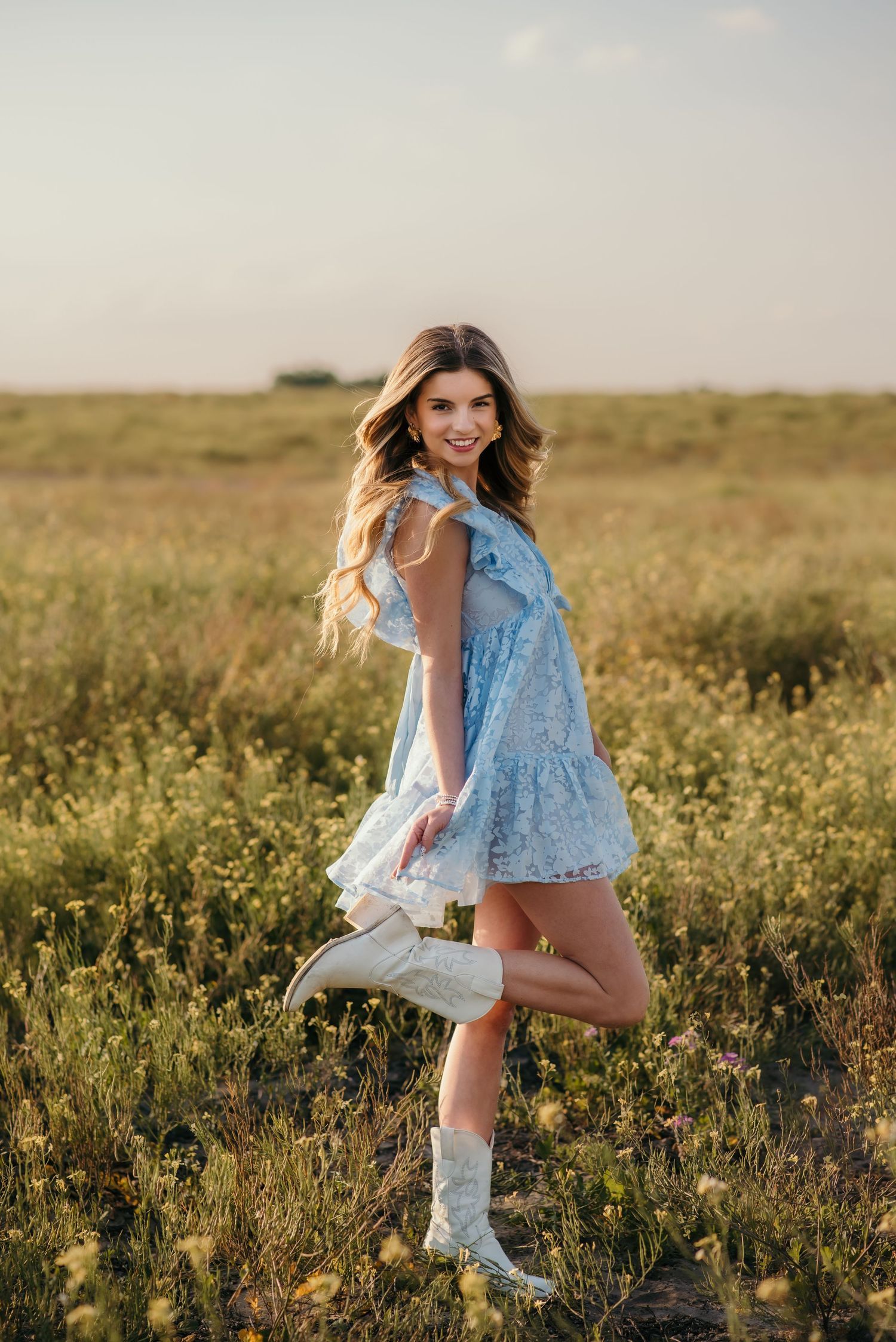 A woman wearing cowboy boots and a blue dress poses playfully in a wildflower field.