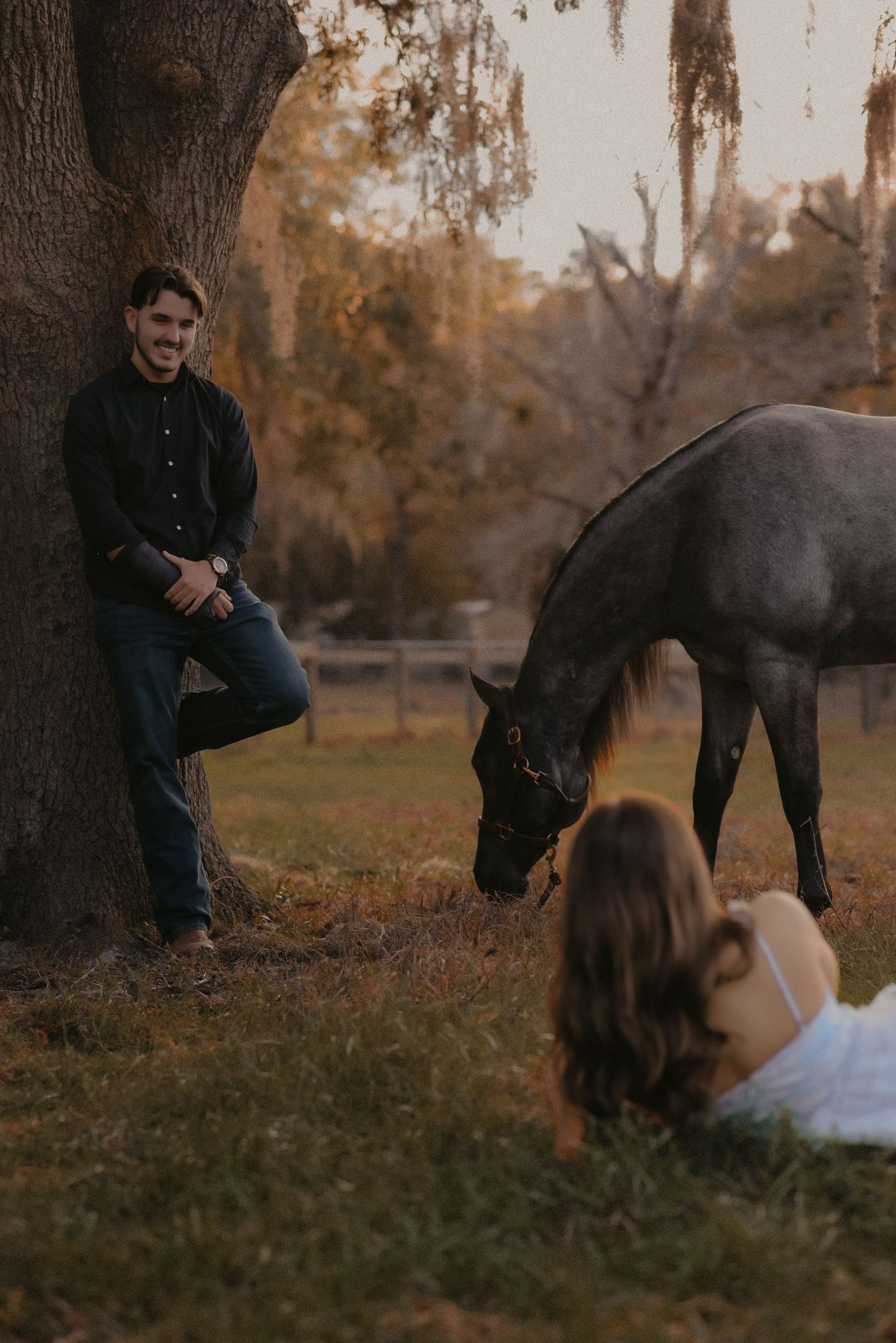 A couple relaxes under autumn trees in a park during golden hour as fallen leaves scatter around them.