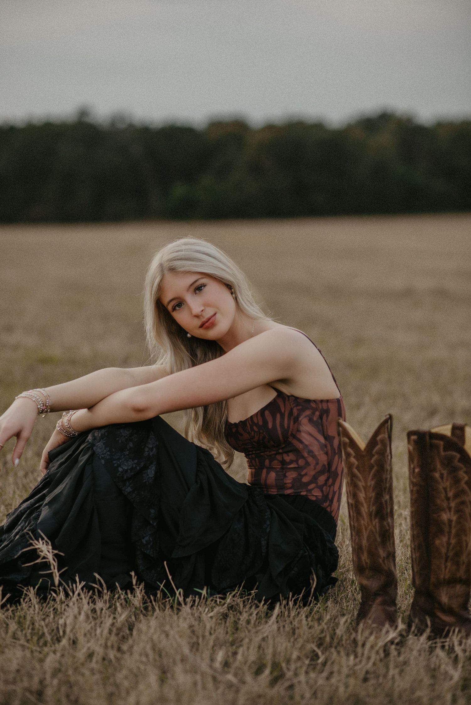 A rustic outdoor portrait in a field features a woman in a dark dress sitting beside brown cowboy boots at sunset.